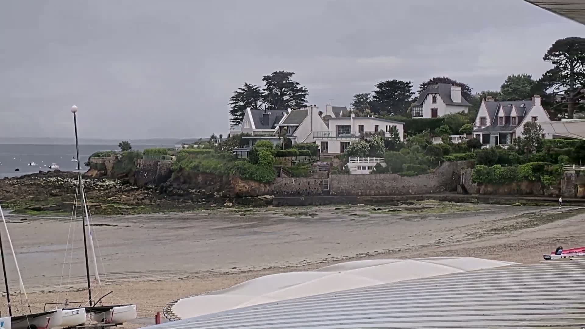 A sandy beach with sailboats and a few houses on a rocky outcrop overlooking a calm, gray sea under an overcast sky.