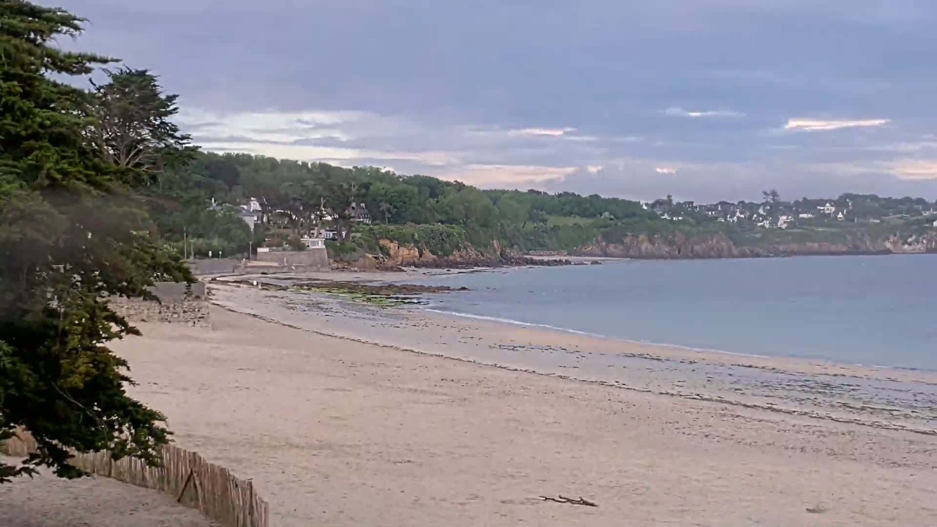 A sandy beach stretches to a calm sea under a partly cloudy sky, backed by a green, tree-covered cliff with houses visible in the distance.