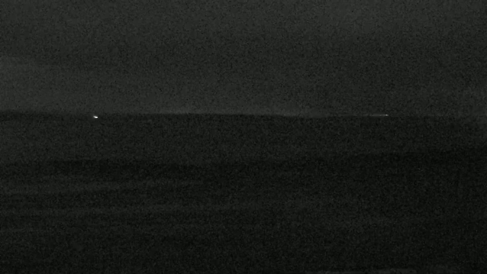 A dark, nighttime image shows a mostly featureless dark body of water with a few faint lights visible on the horizon.