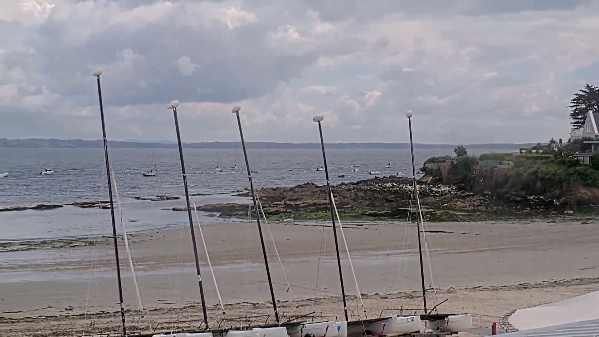 Several sailboats are beached on a sandy shore near dark rocks and calm water under a cloudy sky.