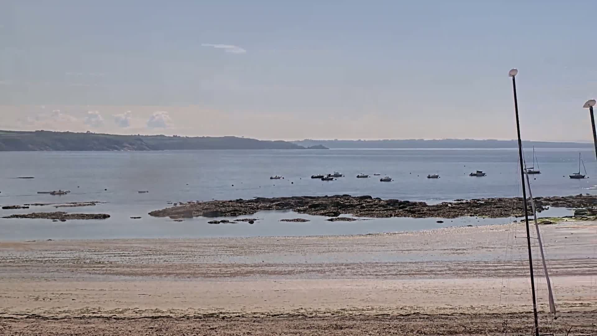 A sandy beach stretches to a calm bay with several small boats and rocky outcroppings under a mostly clear sky.