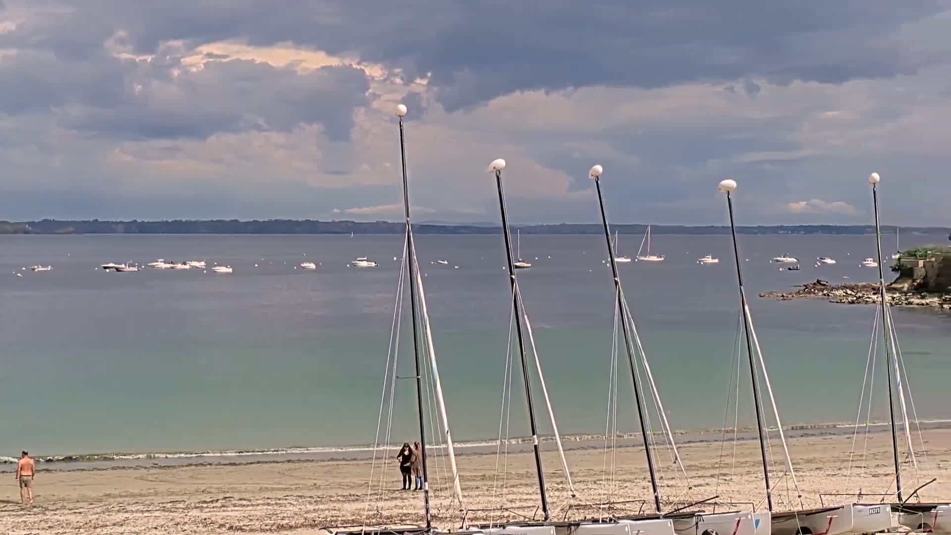 A sandy beach with several sailboats on the shore, a calm body of water with many boats in the distance under a cloudy sky.