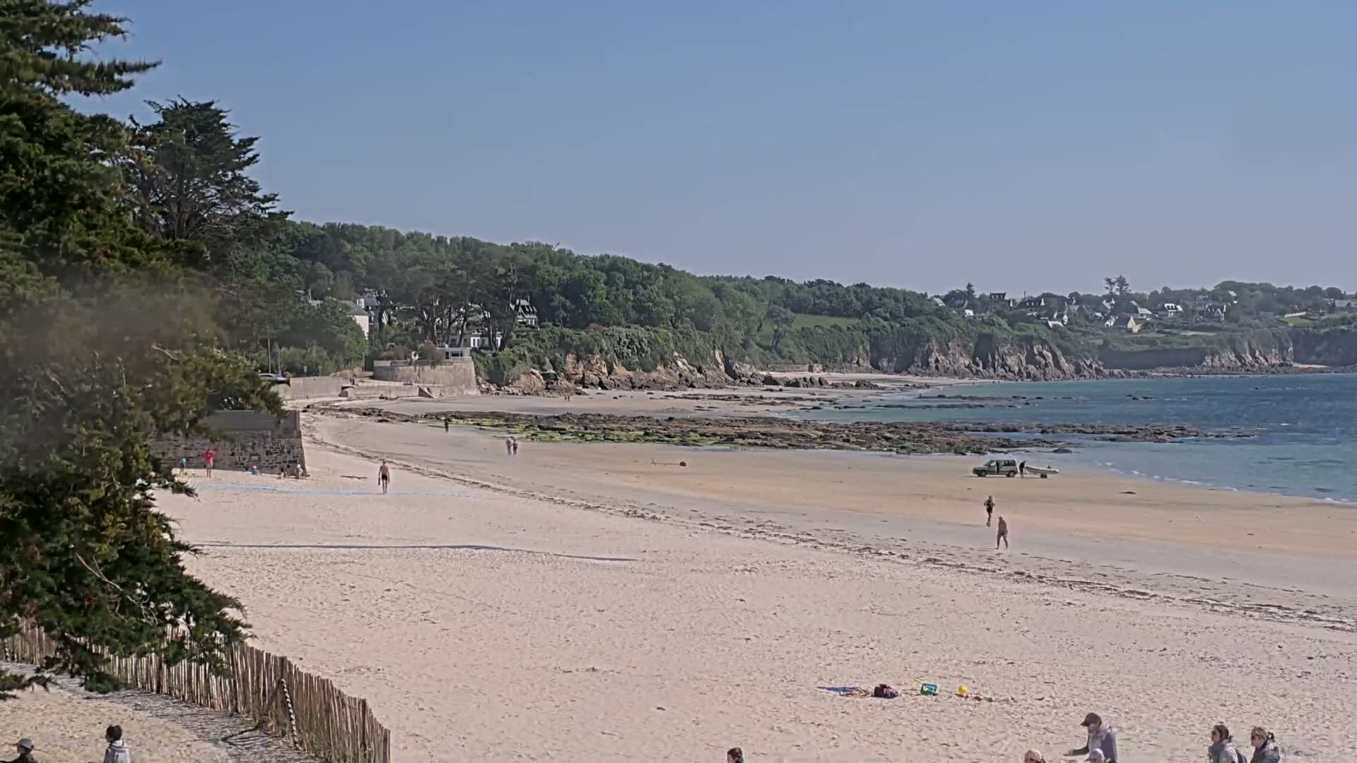 A sandy beach stretches out to a calm ocean under a clear blue sky, with a few people scattered along the shore and a vehicle near the water's edge.
