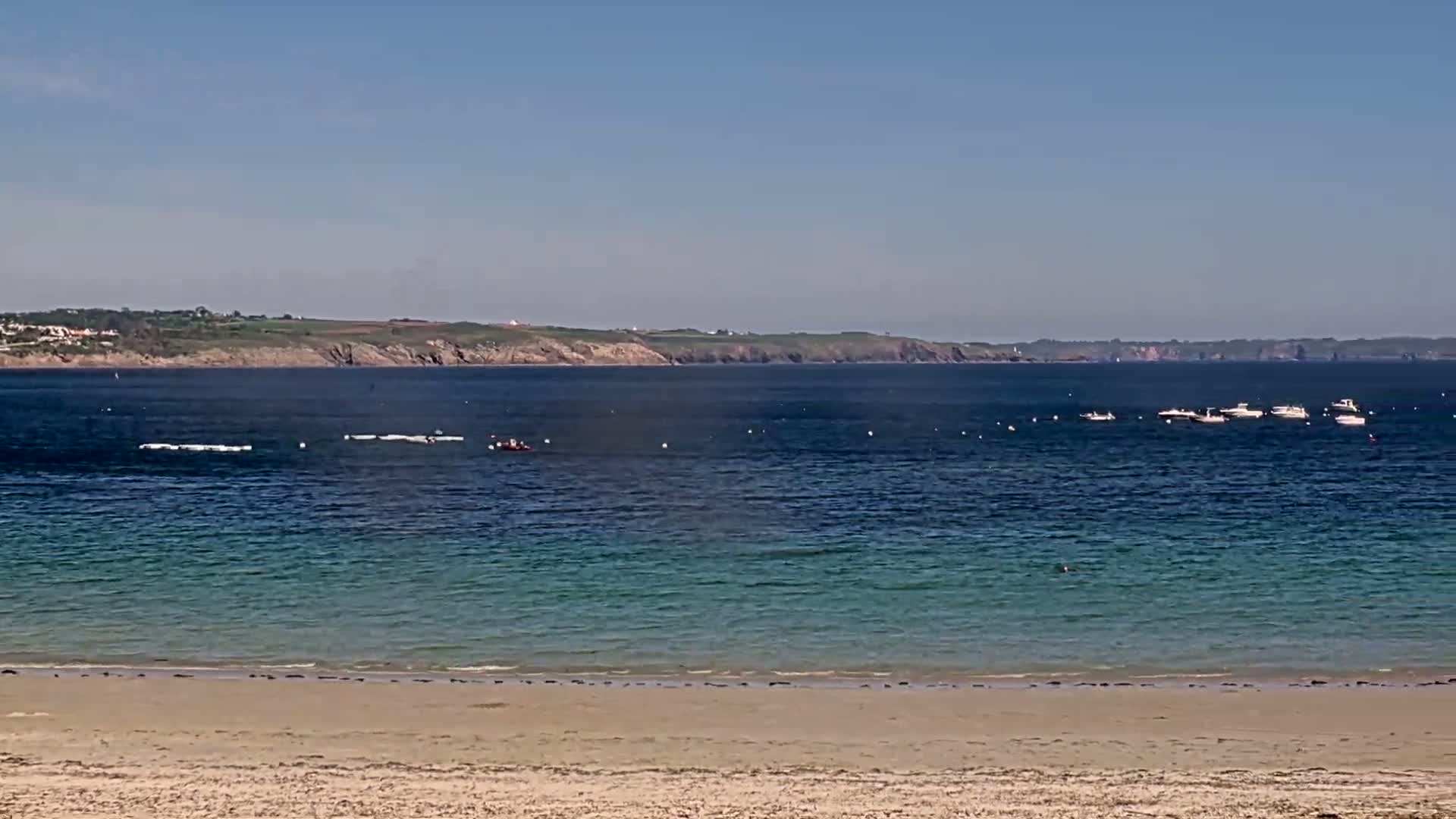A sandy beach stretches to a calm, blue-green ocean where several small boats are anchored, with a distant coastline visible under a clear sky.