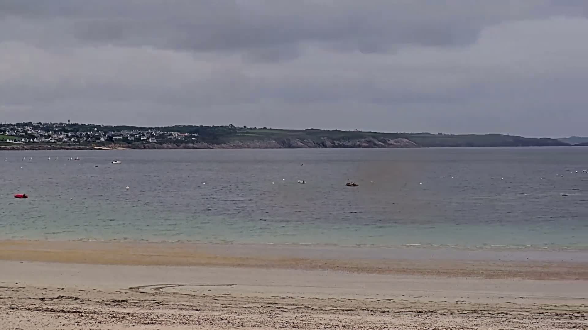 A sandy beach stretches to a calm, pale-green ocean under an overcast sky, with a distant town and coastline visible.