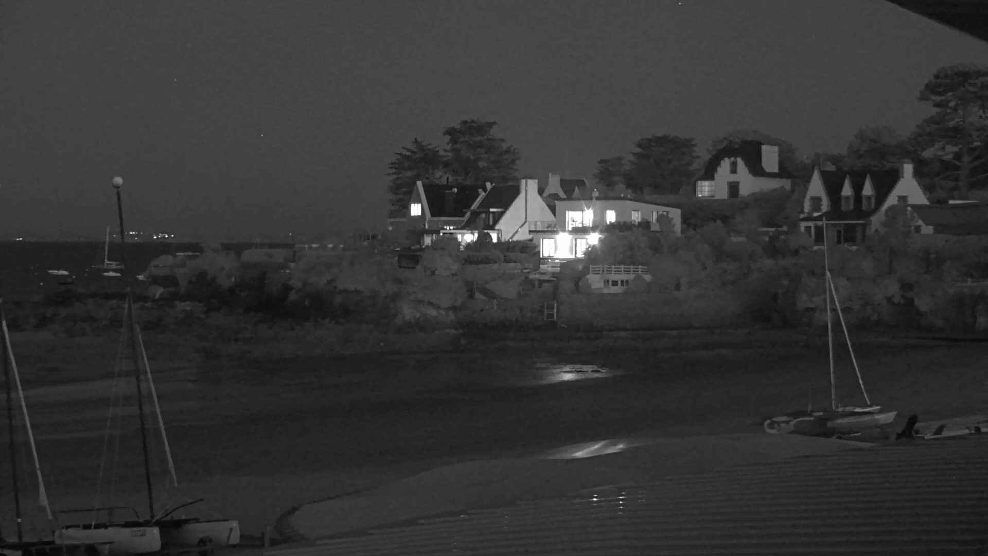 At night, several houses with lights on are situated near a body of water where sailboats are moored.