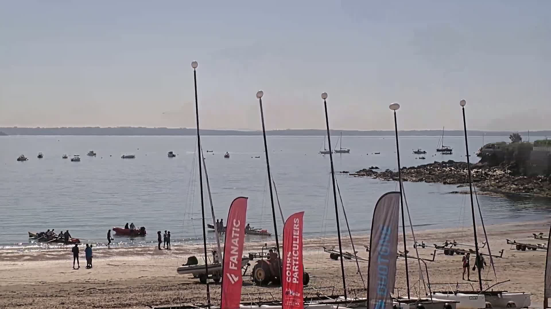 A sandy beach with several sailboats on the water and some people on the shore on a sunny day.
