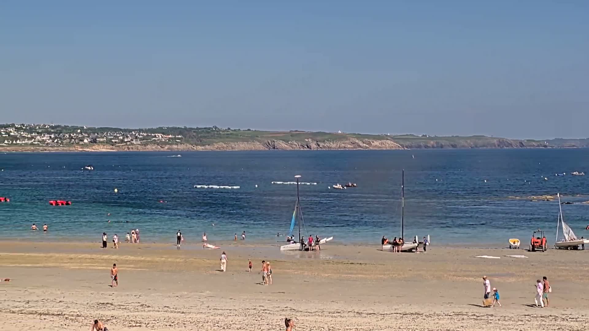 A sandy beach with people swimming and sunbathing, several sailboats beached nearby, and a calm ocean under a clear blue sky.
