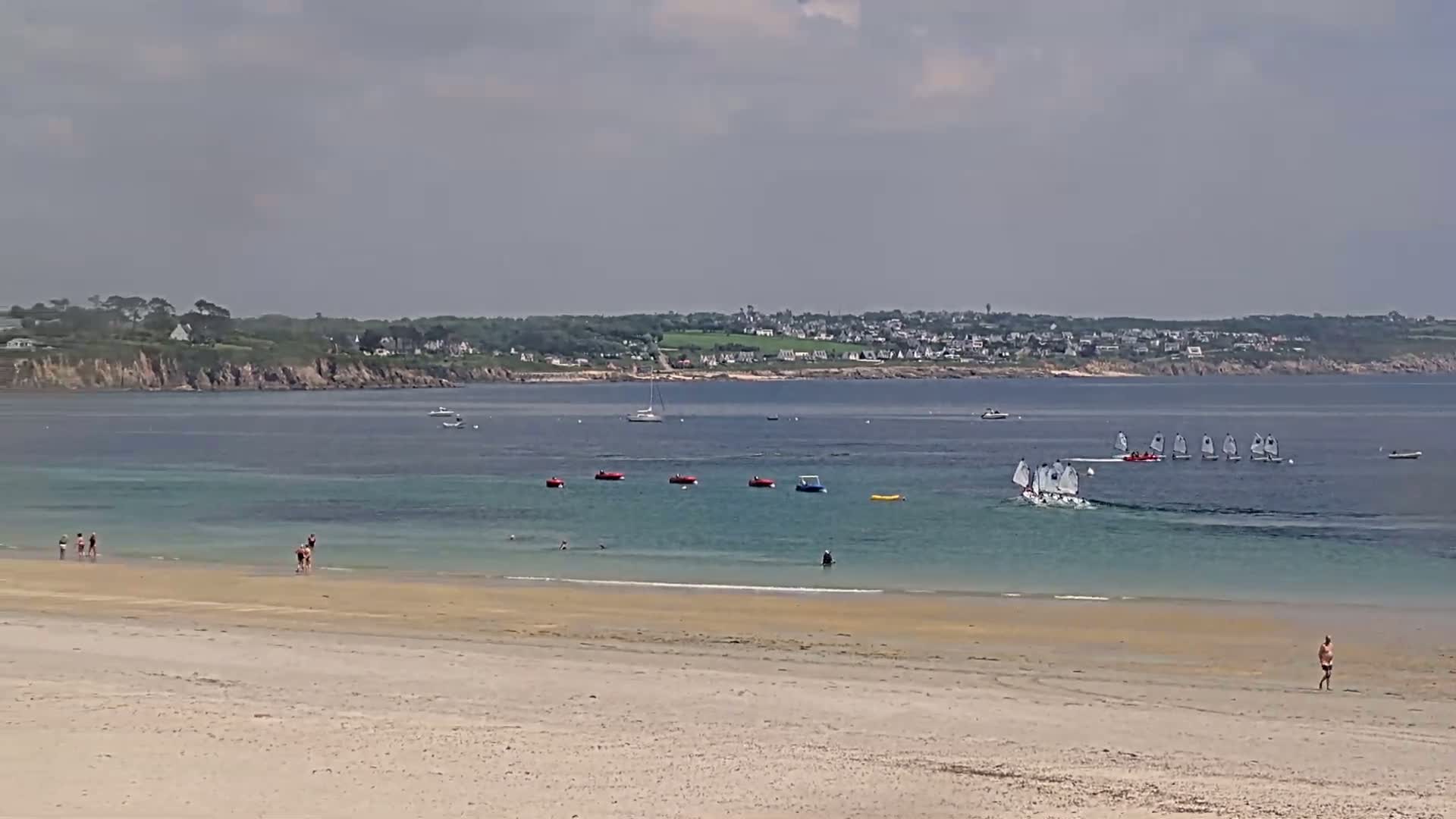 A sandy beach with a few people on it, overlooks calm, shallow turquoise water where several sailboats and small floating objects are visible, and a distant town is seen across a bay under a cloudy sky.