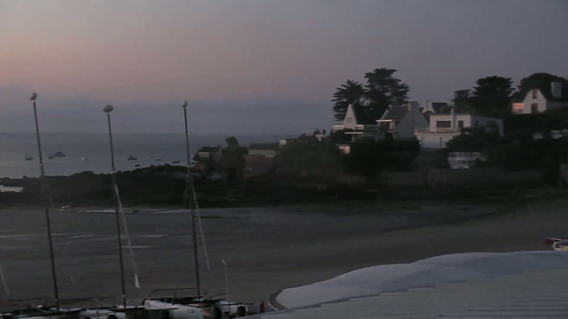 Several sailboats are moored on a calm, dark beach at dusk, with houses and trees visible on a nearby hill under a dusky sky.