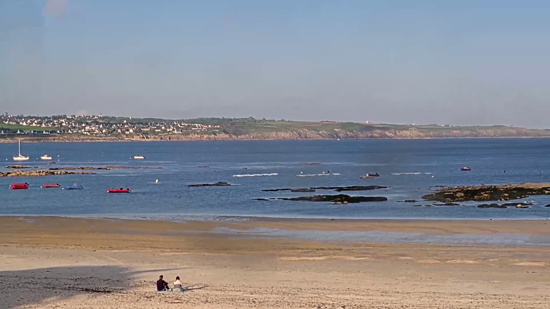 A couple sits on a sandy beach overlooking a calm bay with several small boats and rocky outcroppings, under a clear blue sky.