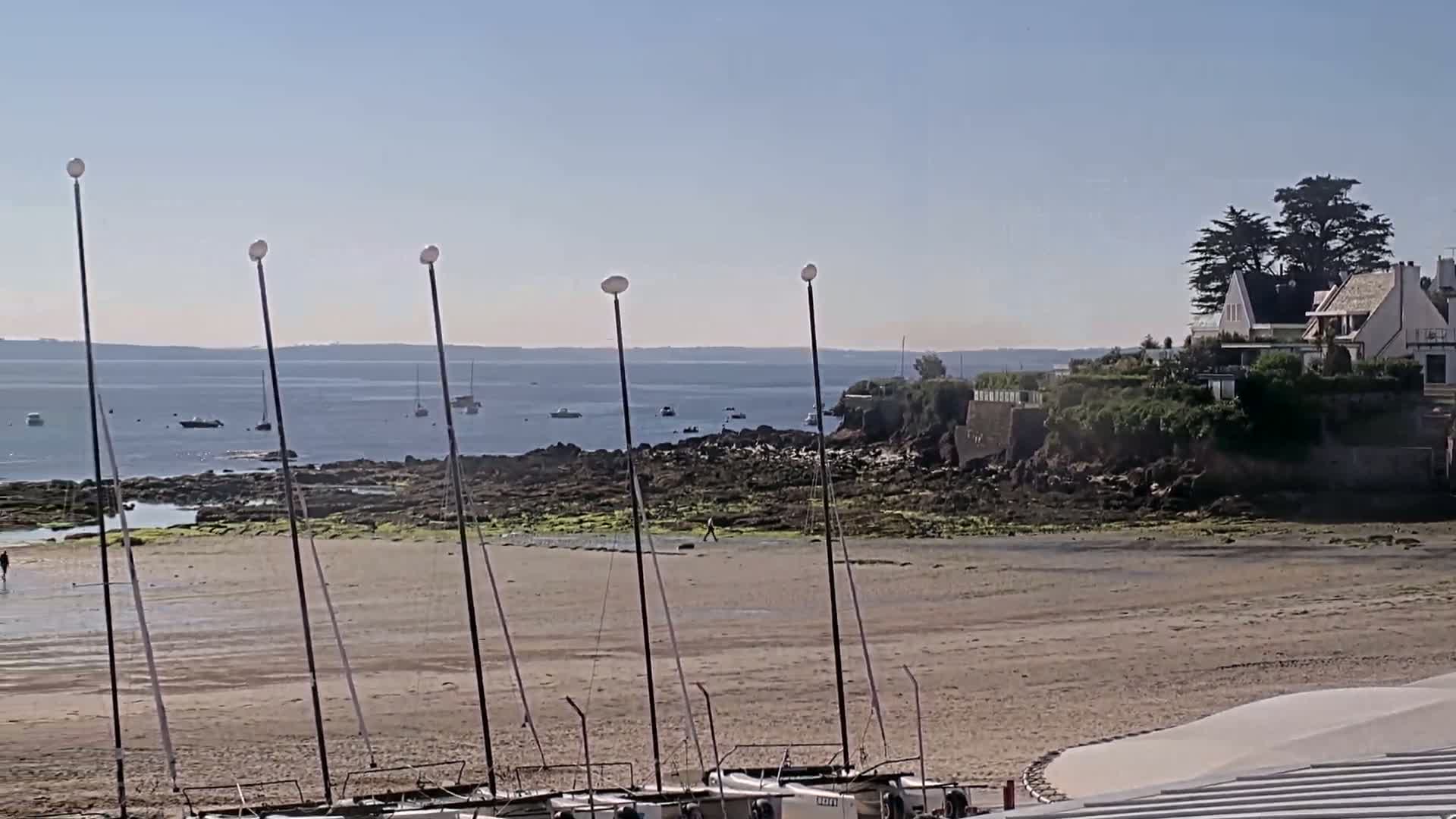 A sandy beach with several sailboats beached on it, a rocky shoreline, calm water with a few boats visible on the horizon, and a house on a cliff overlooking the water on a sunny day.