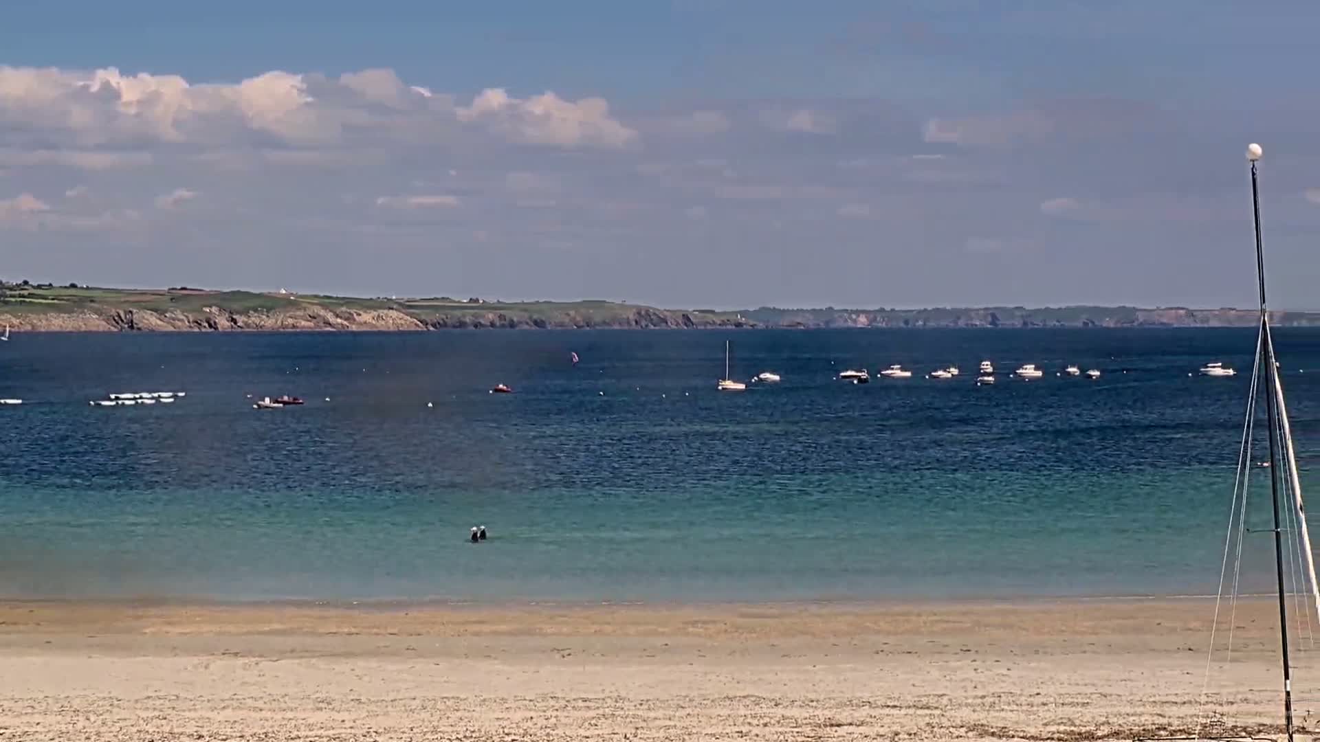 A sandy beach stretches to a calm, blue-green sea where several boats are anchored, under a partly cloudy sky.