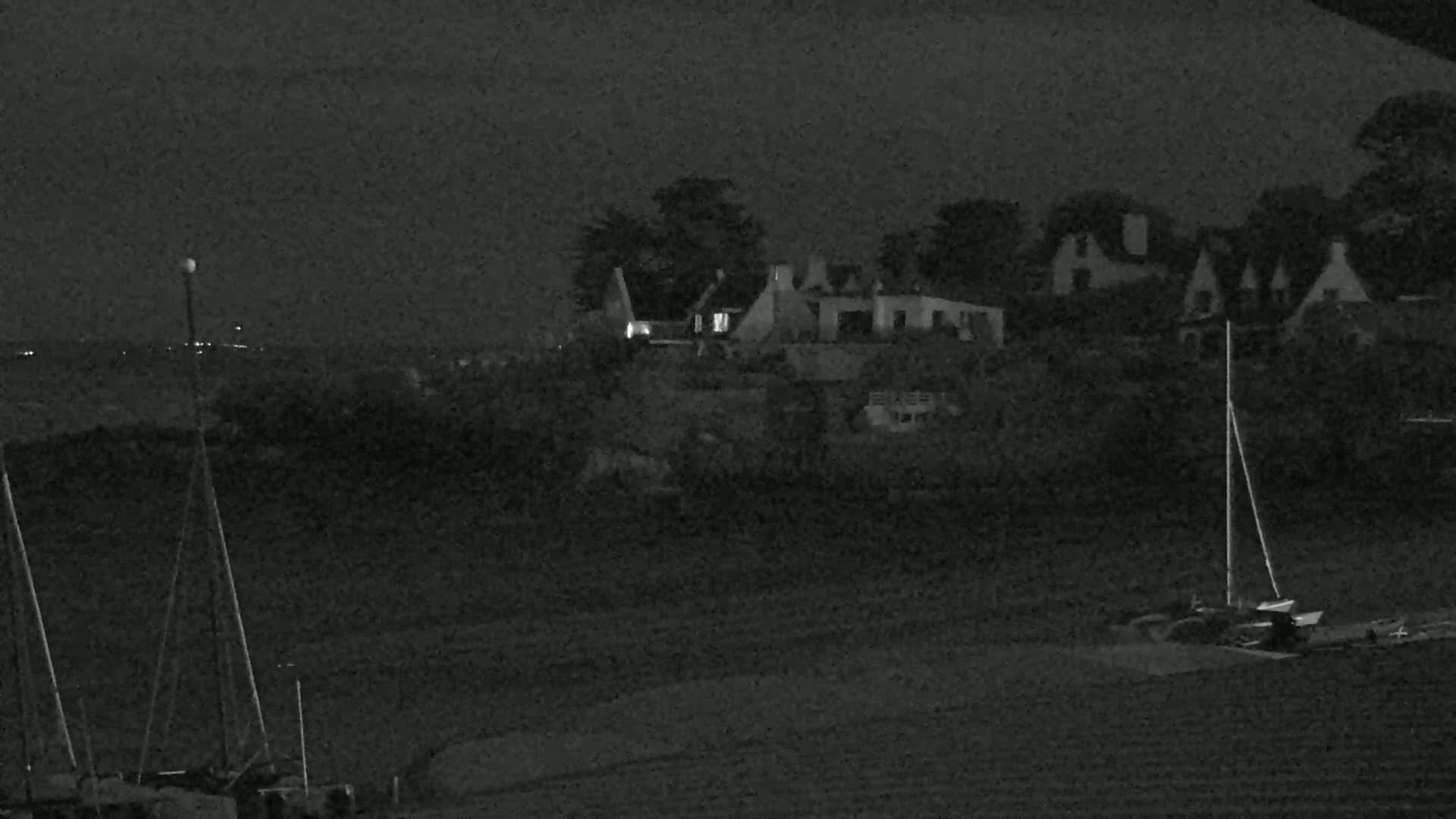 A nighttime, low-light image shows several buildings near a body of water, with sailboats moored in the foreground.