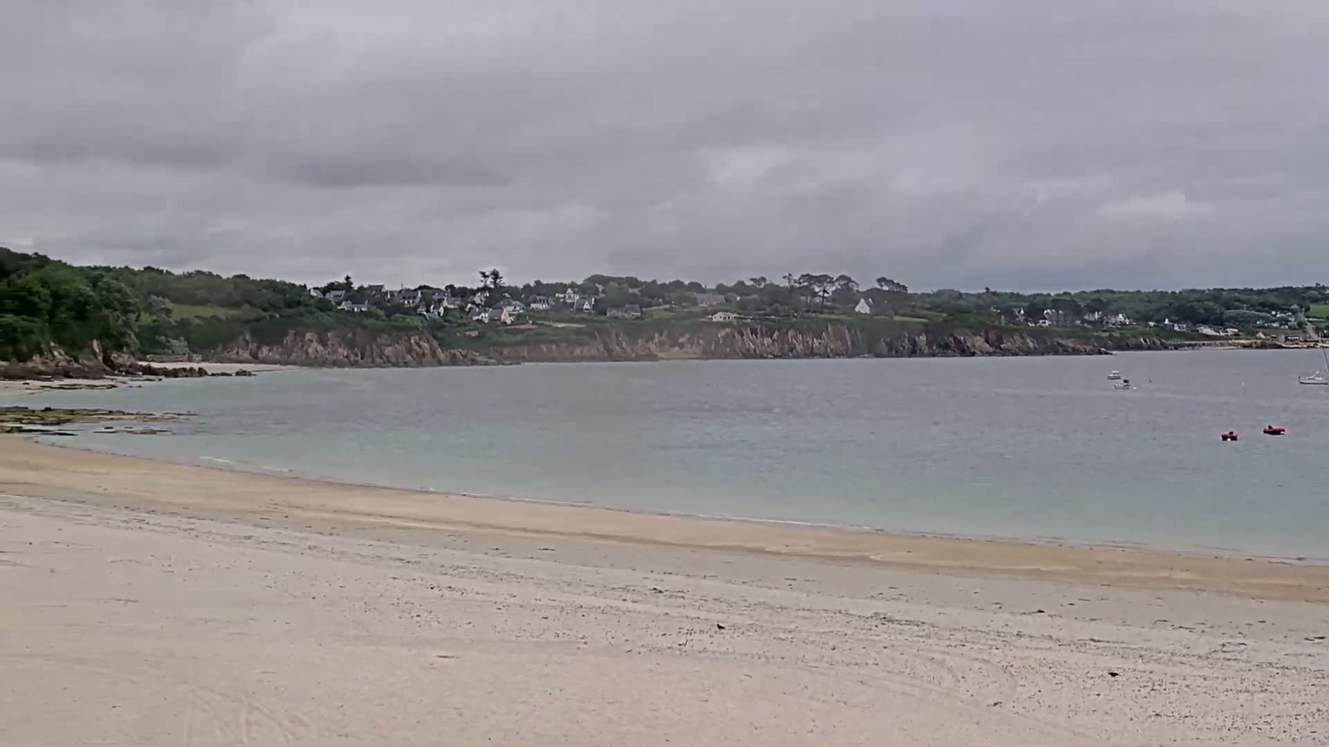 A sandy beach stretches out to a calm, light-blue bay under an overcast sky, with a line of houses visible on a distant, green hillside.