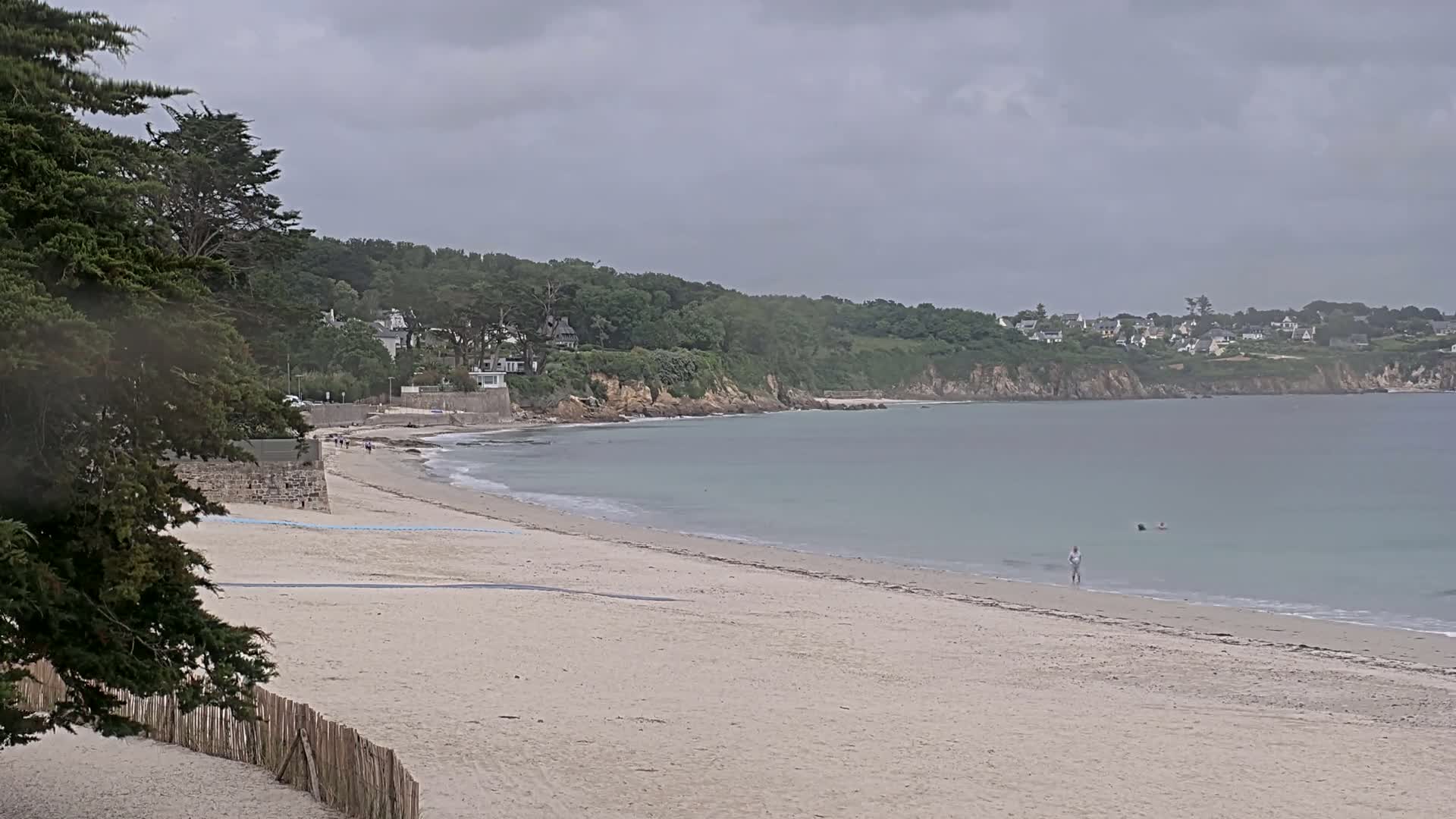 A sandy beach with calm, light-blue water stretches out under an overcast sky, backed by a lush green hillside with a few buildings visible in the distance.
