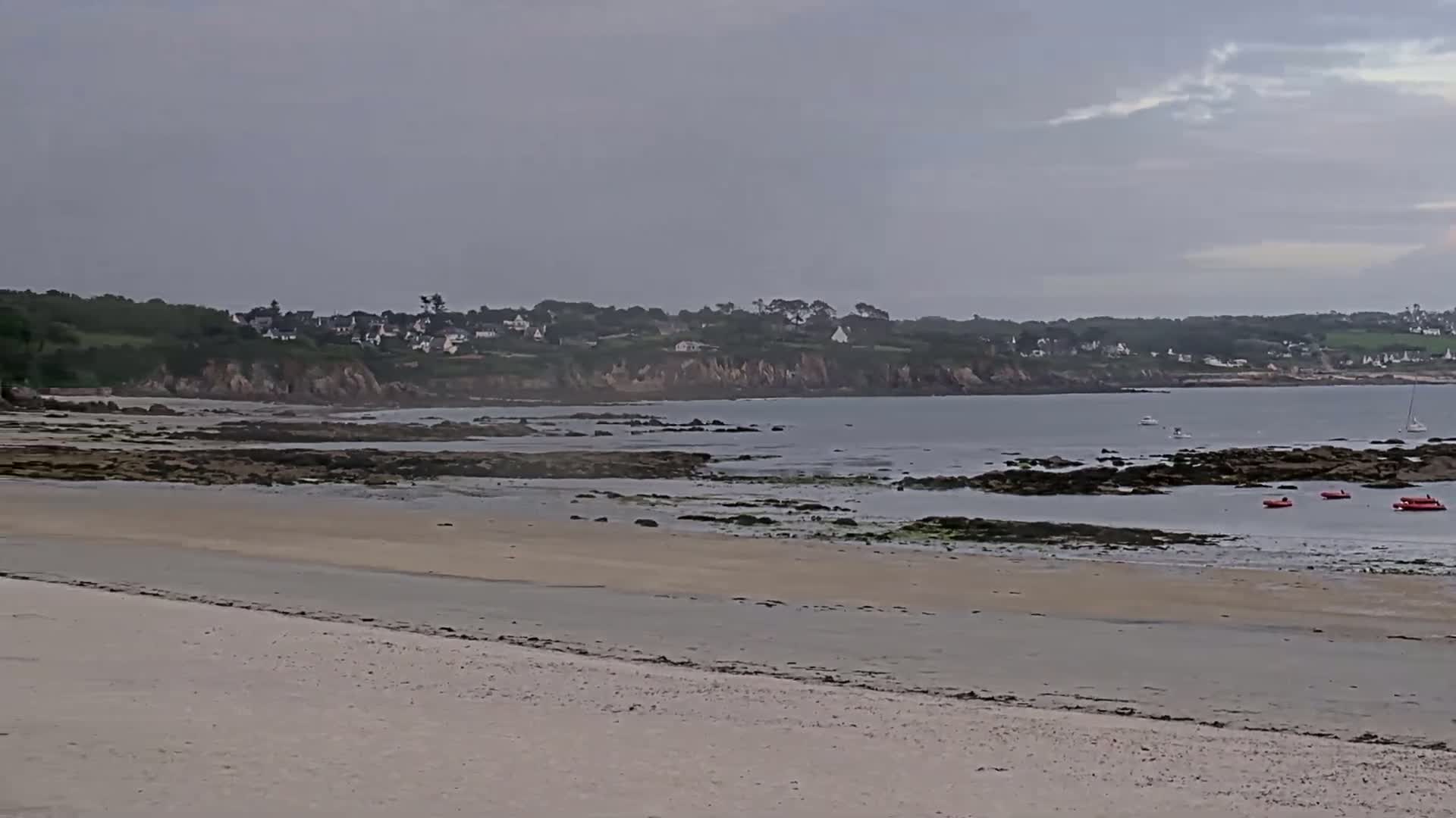 A sandy beach stretches to a calm, gray-sky sea with rocky intertidal zones, small boats, and a distant village nestled against a green hillside.