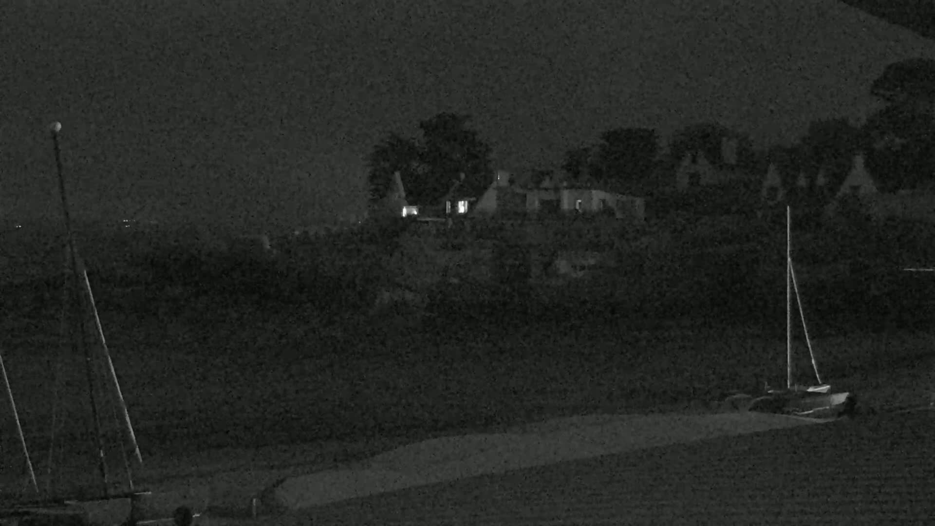 At night, two sailboats are moored near a shoreline with a building and trees under a dark sky.
