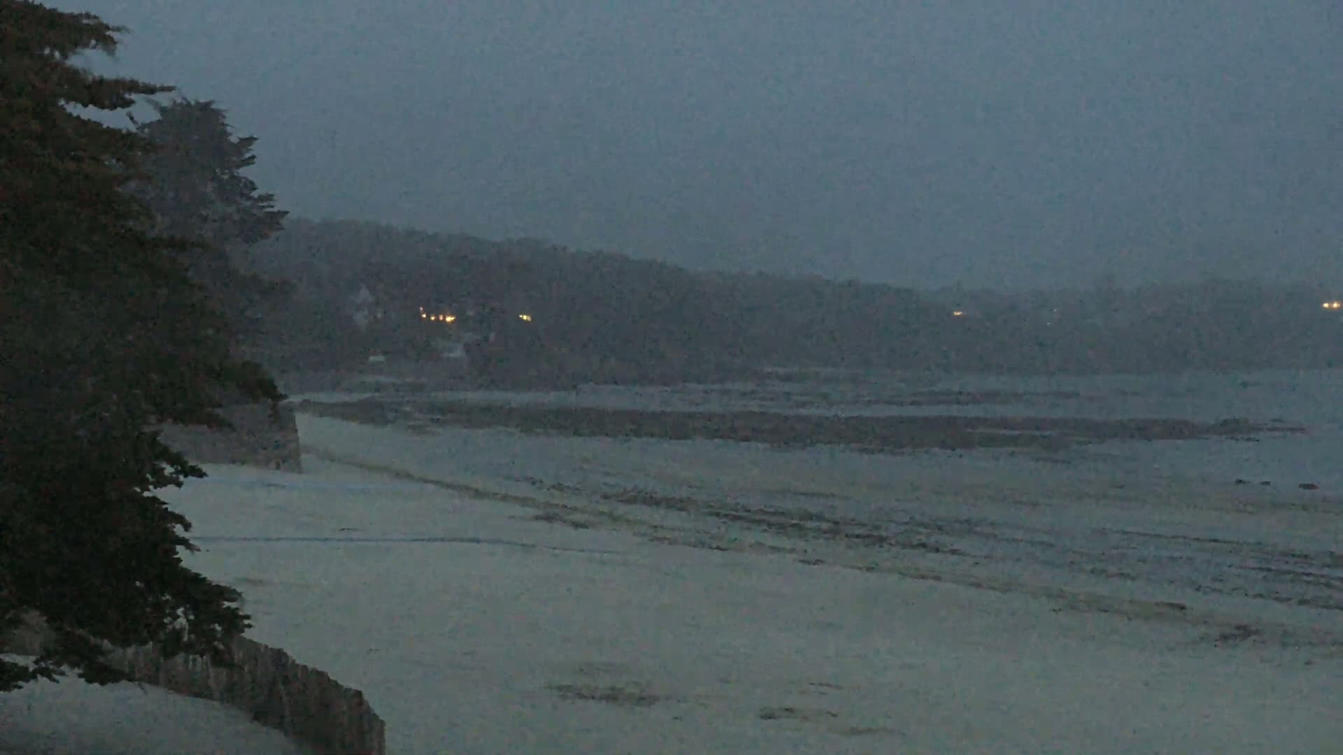 A sandy beach stretches out to a calm, dark ocean under a hazy, overcast sky, with distant lights visible on the shoreline across the water.