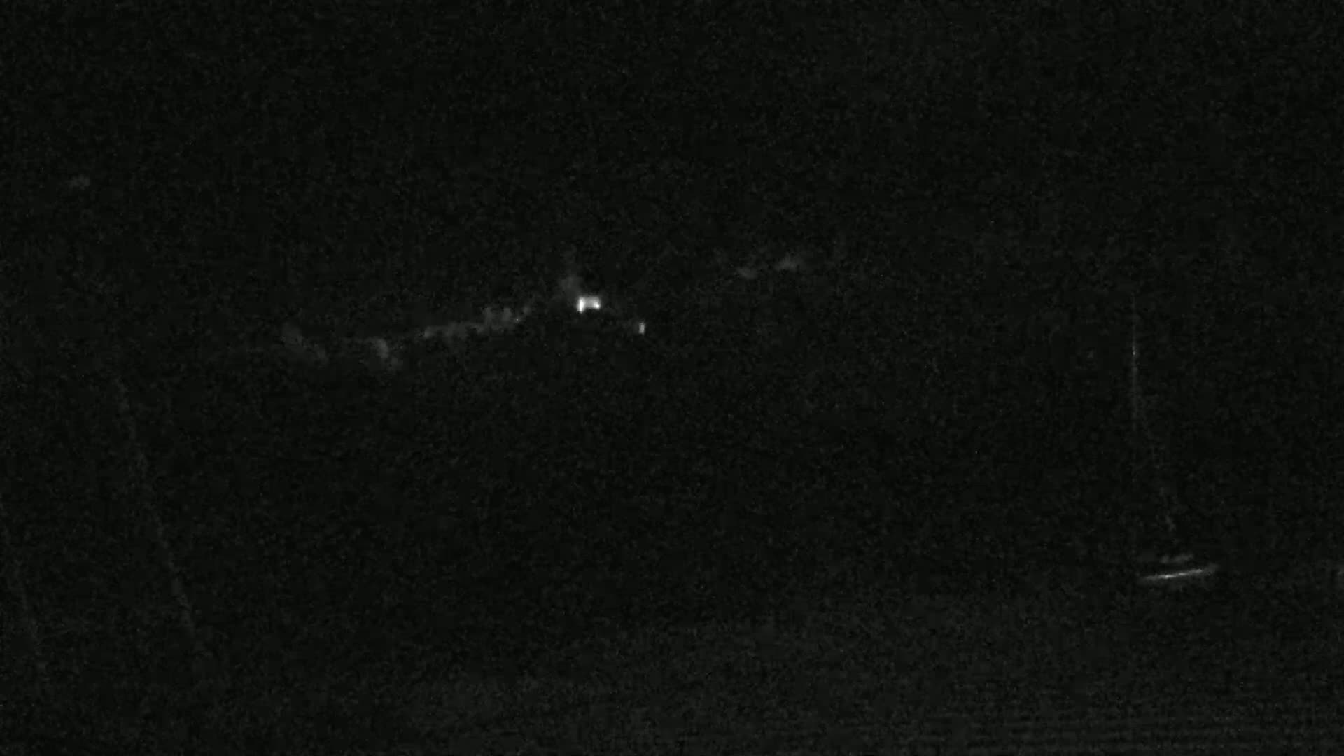 A nighttime, low-light image shows a dark, indistinct landscape with a few faint lights in the distance.