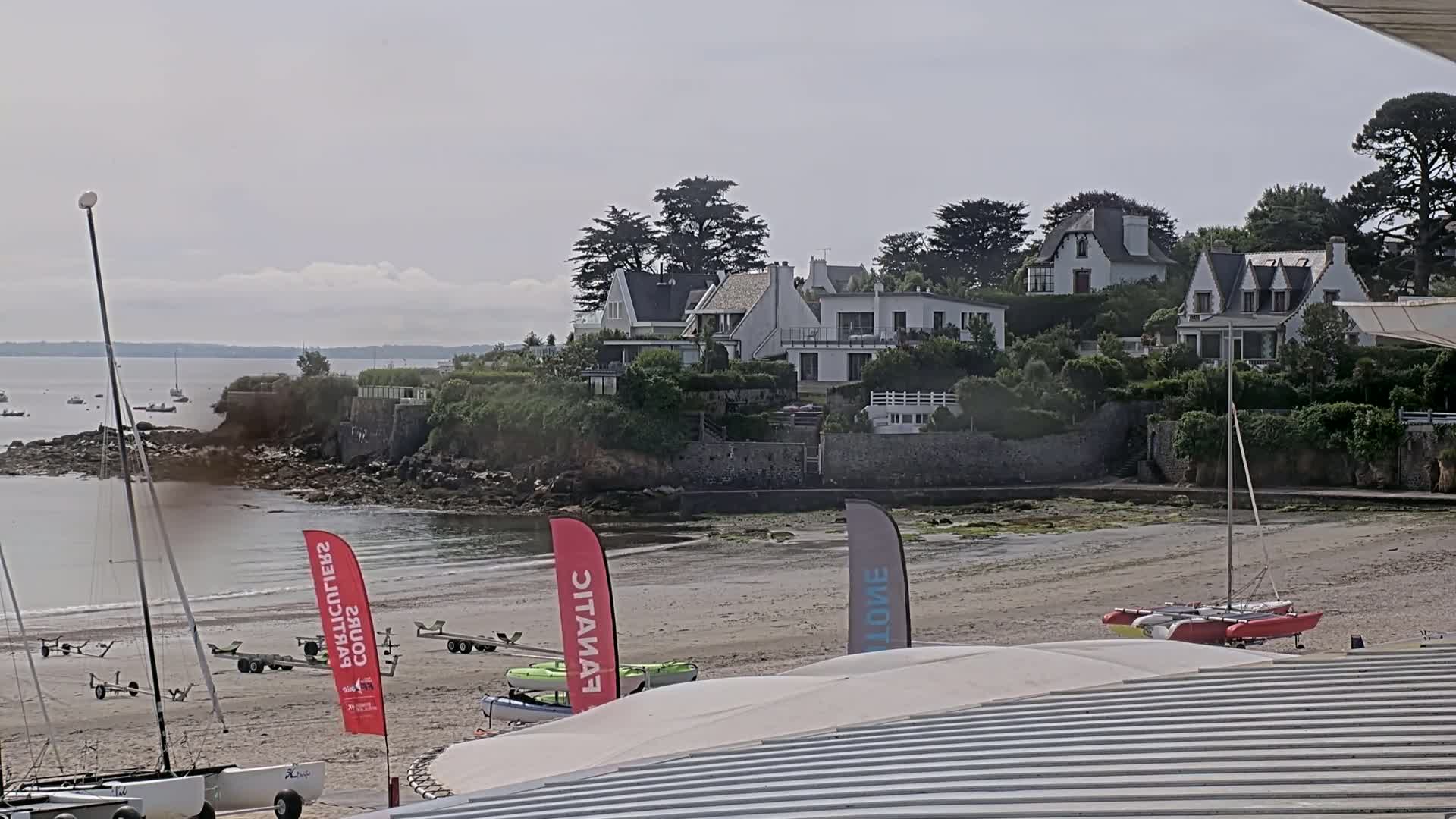 Several sailboats and catamarans are beached on a sandy shore near a cluster of white houses on a slightly overcast day.
