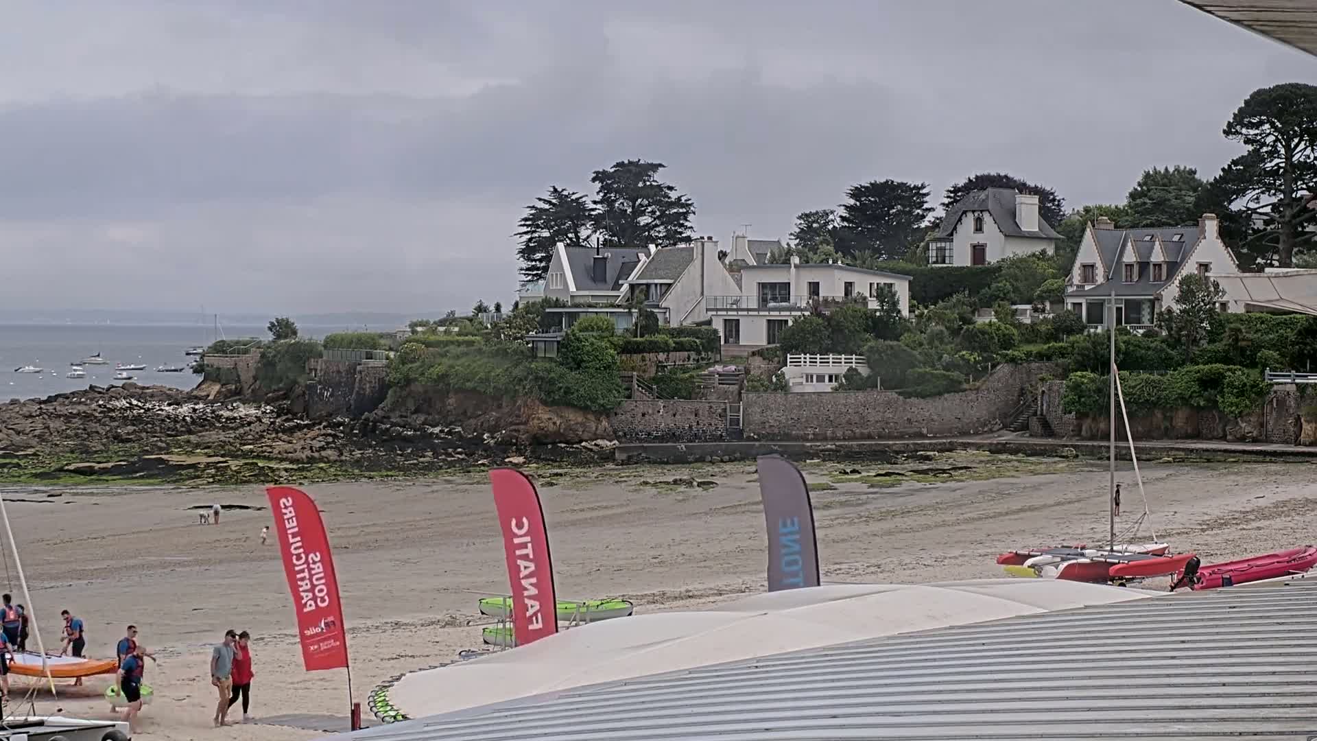 A sandy beach with several sailboats and people walking near a rocky shoreline and a cluster of white houses under an overcast sky.