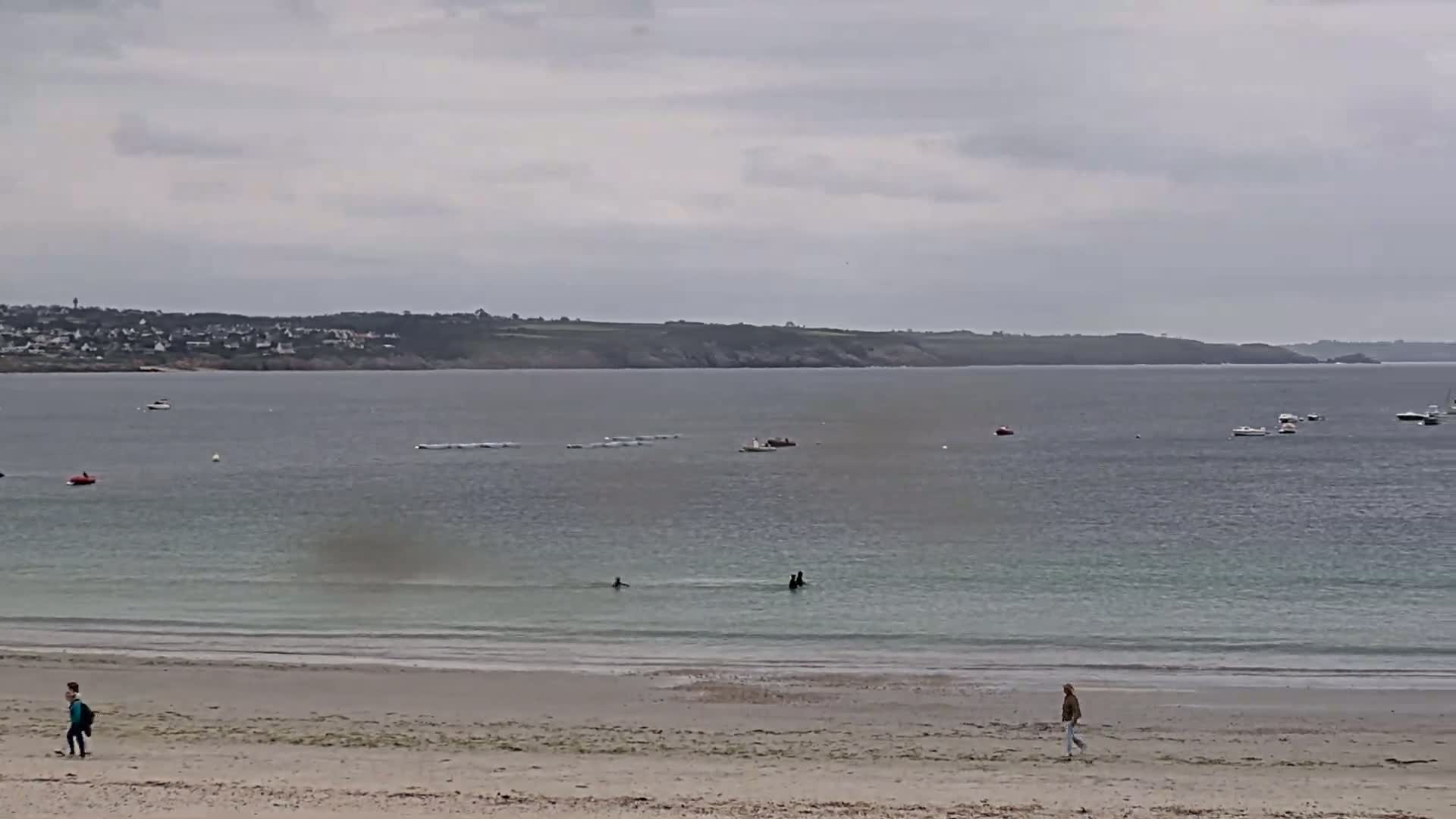 A sandy beach with a few people walking on it, overlooks calm, light-green water with several boats and people swimming further out, under an overcast sky.
