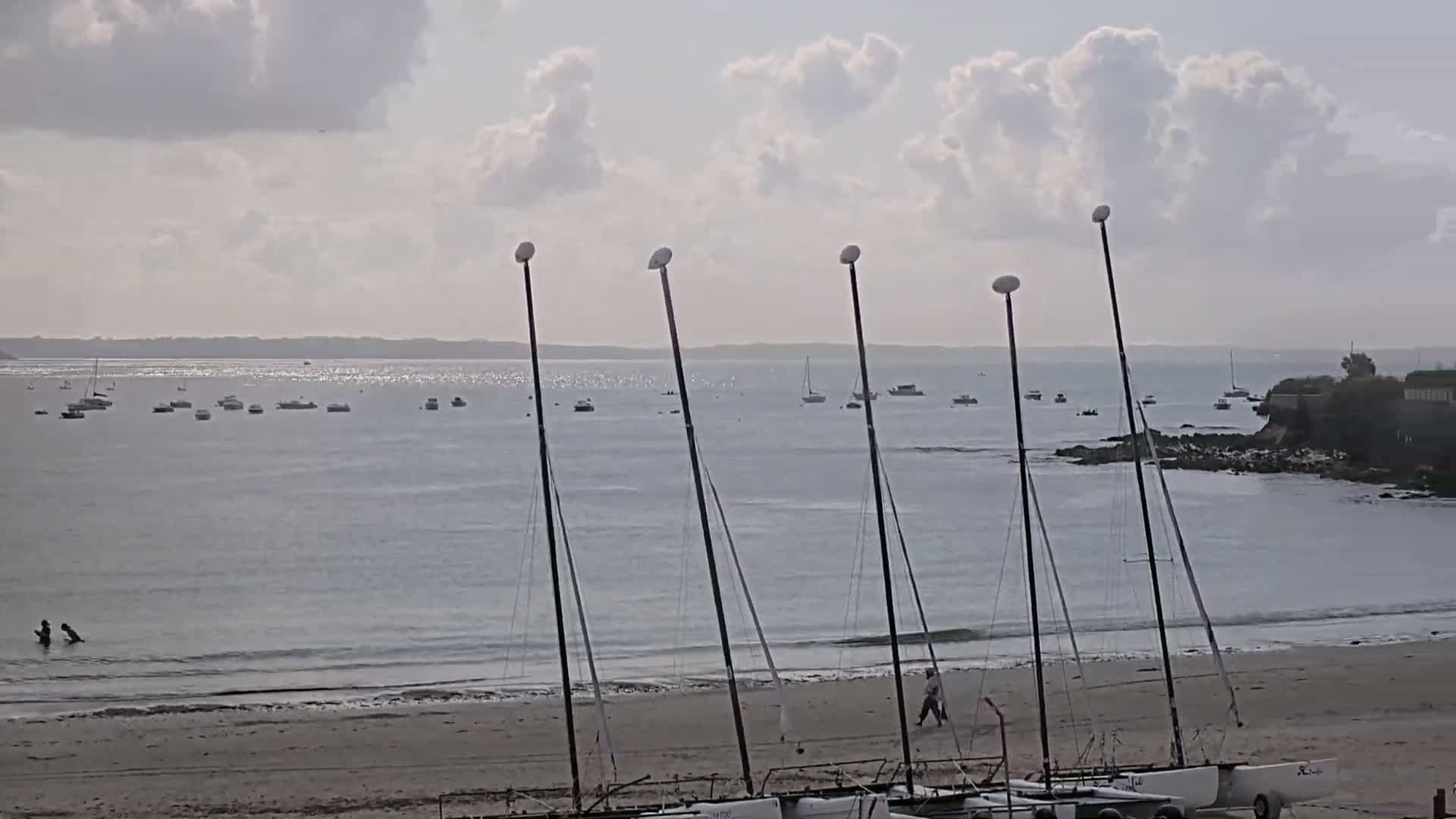Several sailboats are beached on a sandy shore under a partly cloudy sky, with calm water and numerous boats visible in the distance.