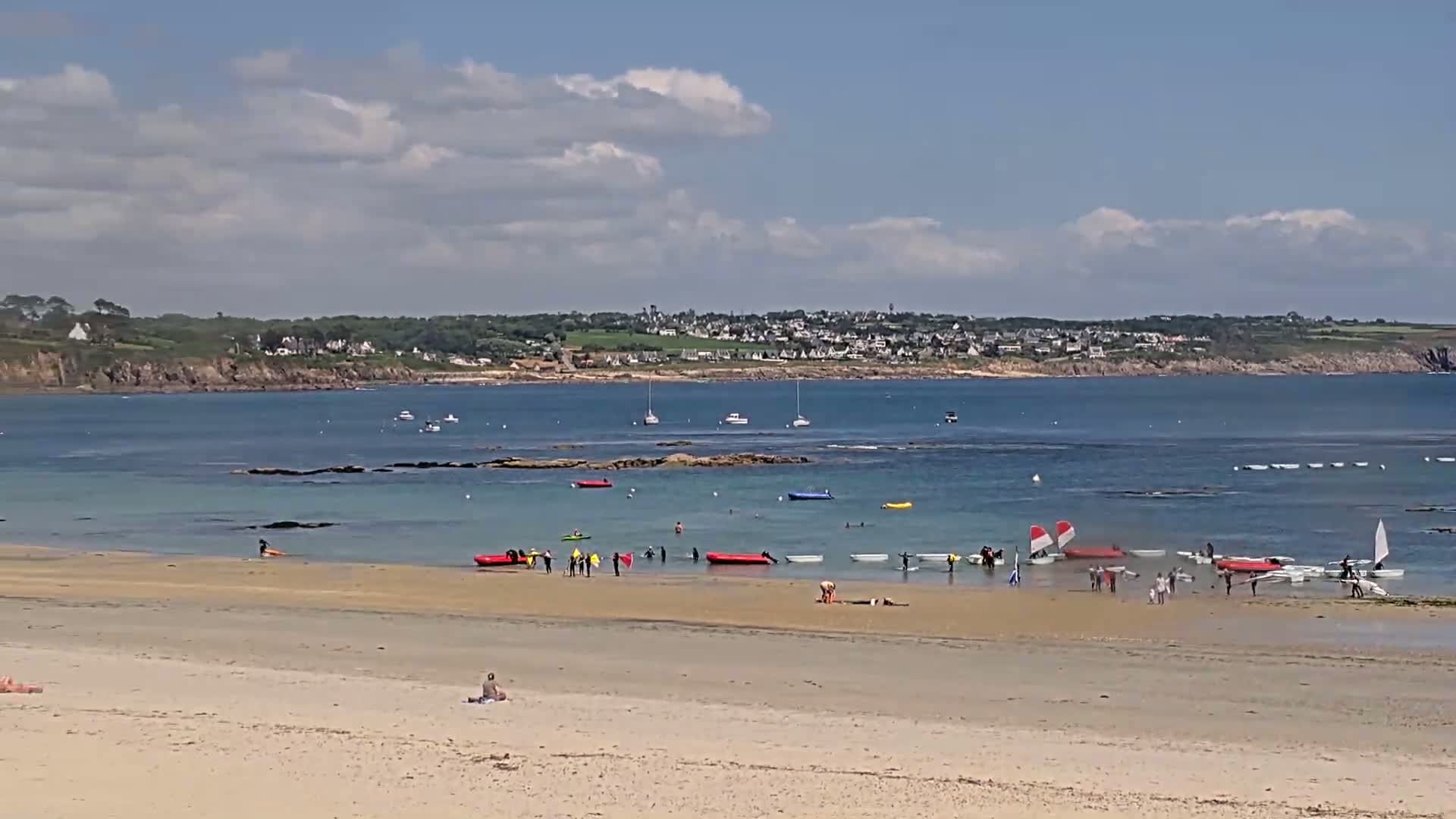 A sandy beach with people sunbathing and participating in watersports, such as kayaking and sailing, under a partly cloudy sky.
