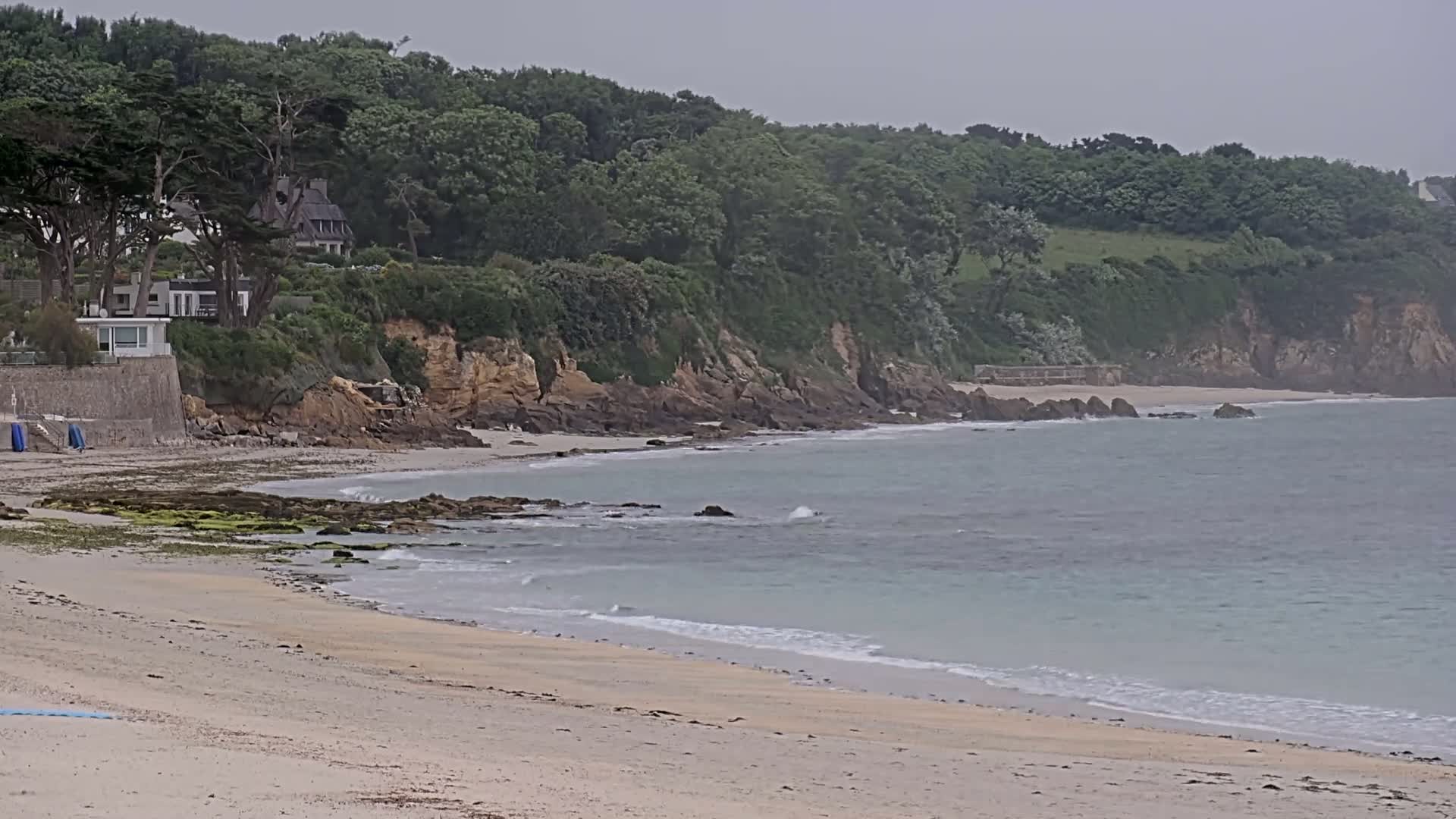 A sandy beach with calm, light-blue water laps gently against the shore, backed by a rocky cliff topped with lush green trees and houses under an overcast sky.