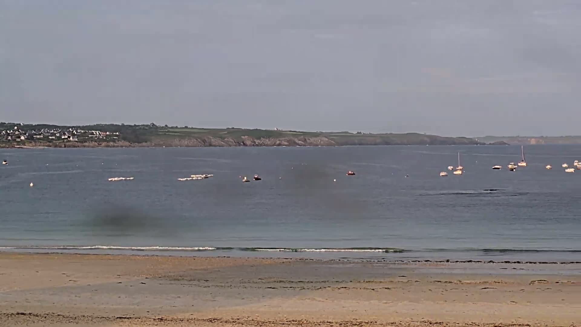 A sandy beach meets a calm sea dotted with small boats and distant land under a hazy sky.