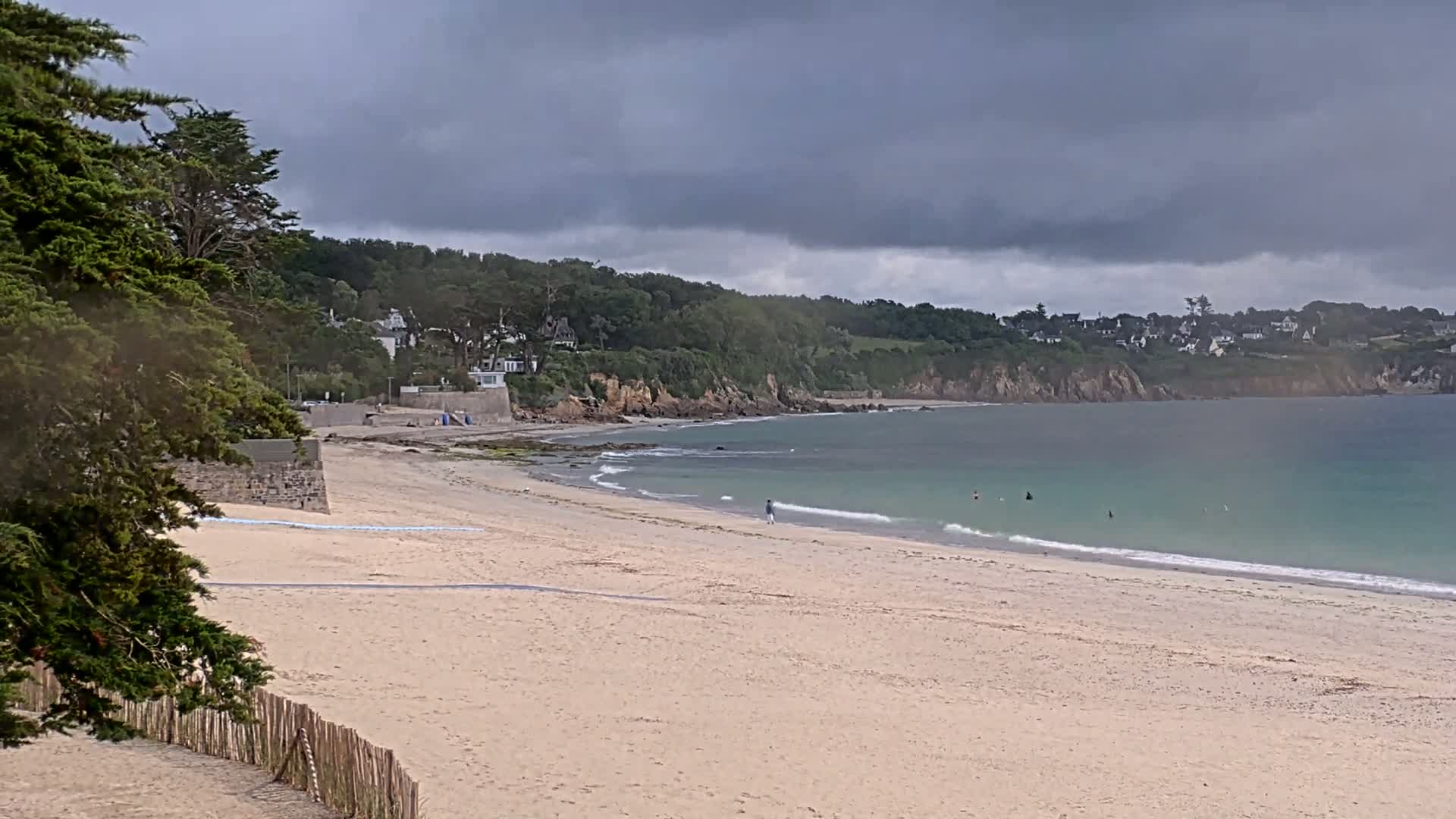 A sandy beach stretches out to a calm, teal ocean under a cloudy sky, backed by a green, hilly coastline with houses scattered along the top.