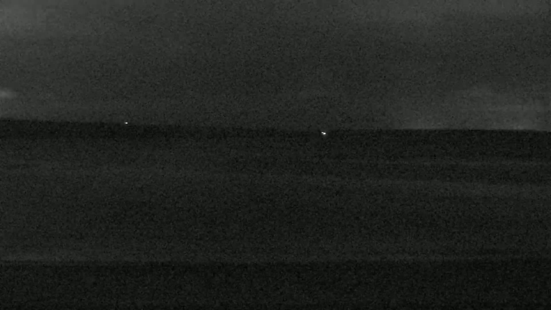 A dark, nighttime scene shows a flat expanse of land or water under a mostly cloudy sky, with two faint lights visible in the distance.
