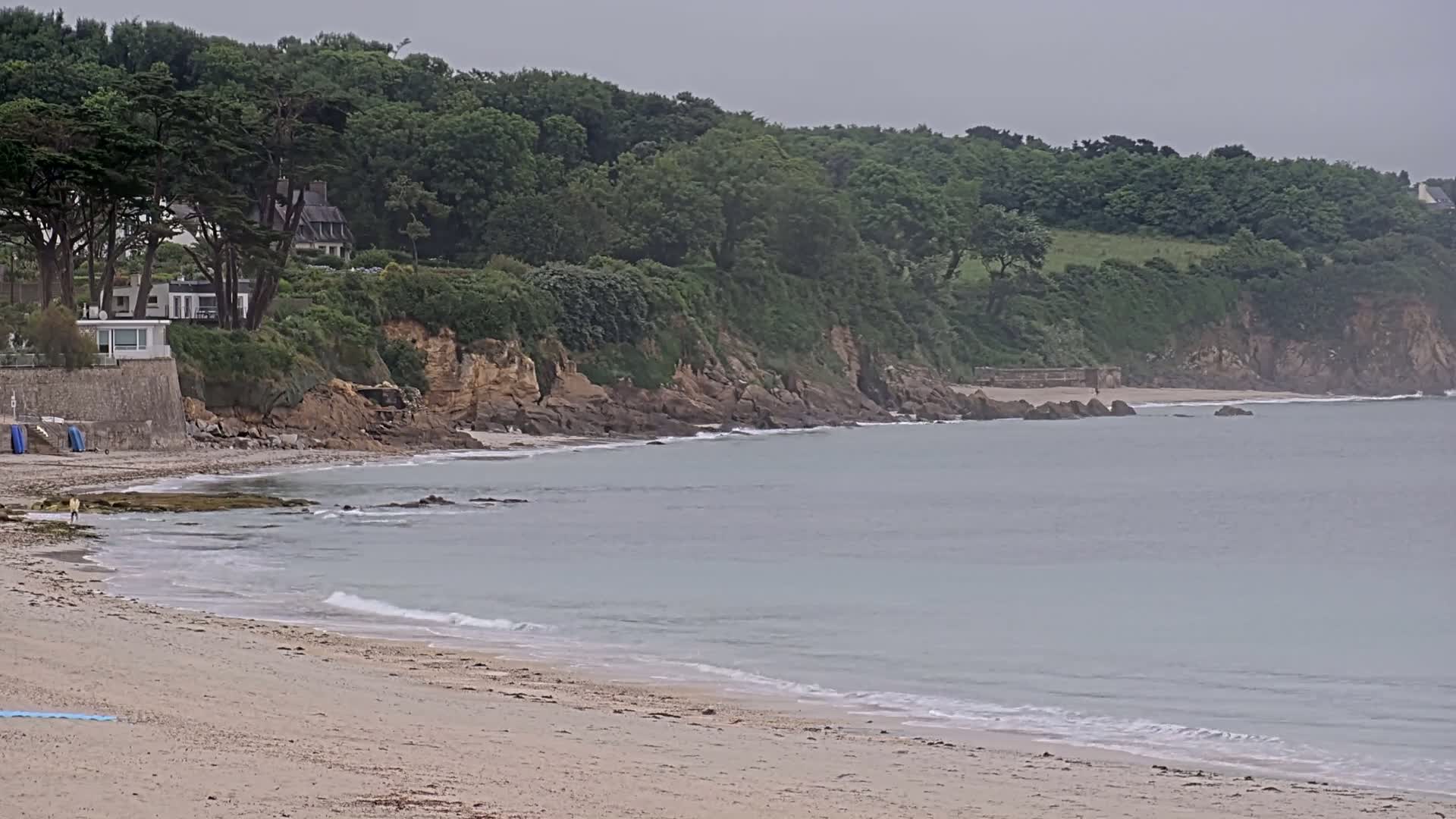 A sandy beach with gentle waves laps at the shore, under an overcast sky, beside a rocky coastline lined with lush green trees and houses.
