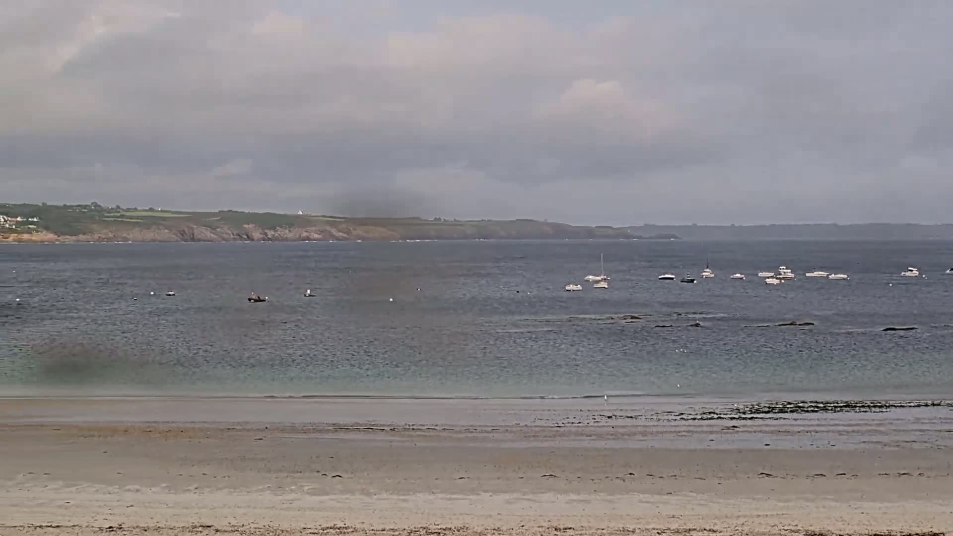 A sandy beach stretches to a calm sea where several small boats are anchored, under a cloudy sky.