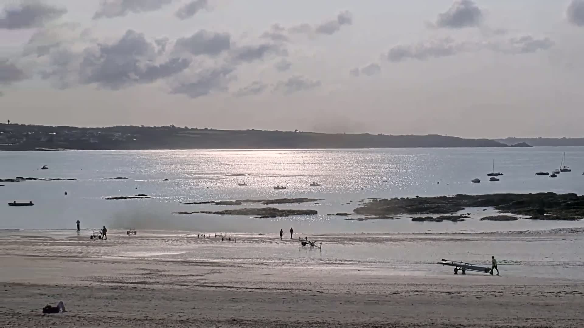 A sandy beach with several people and boats on a calm, glittering sea under a partly cloudy sky.