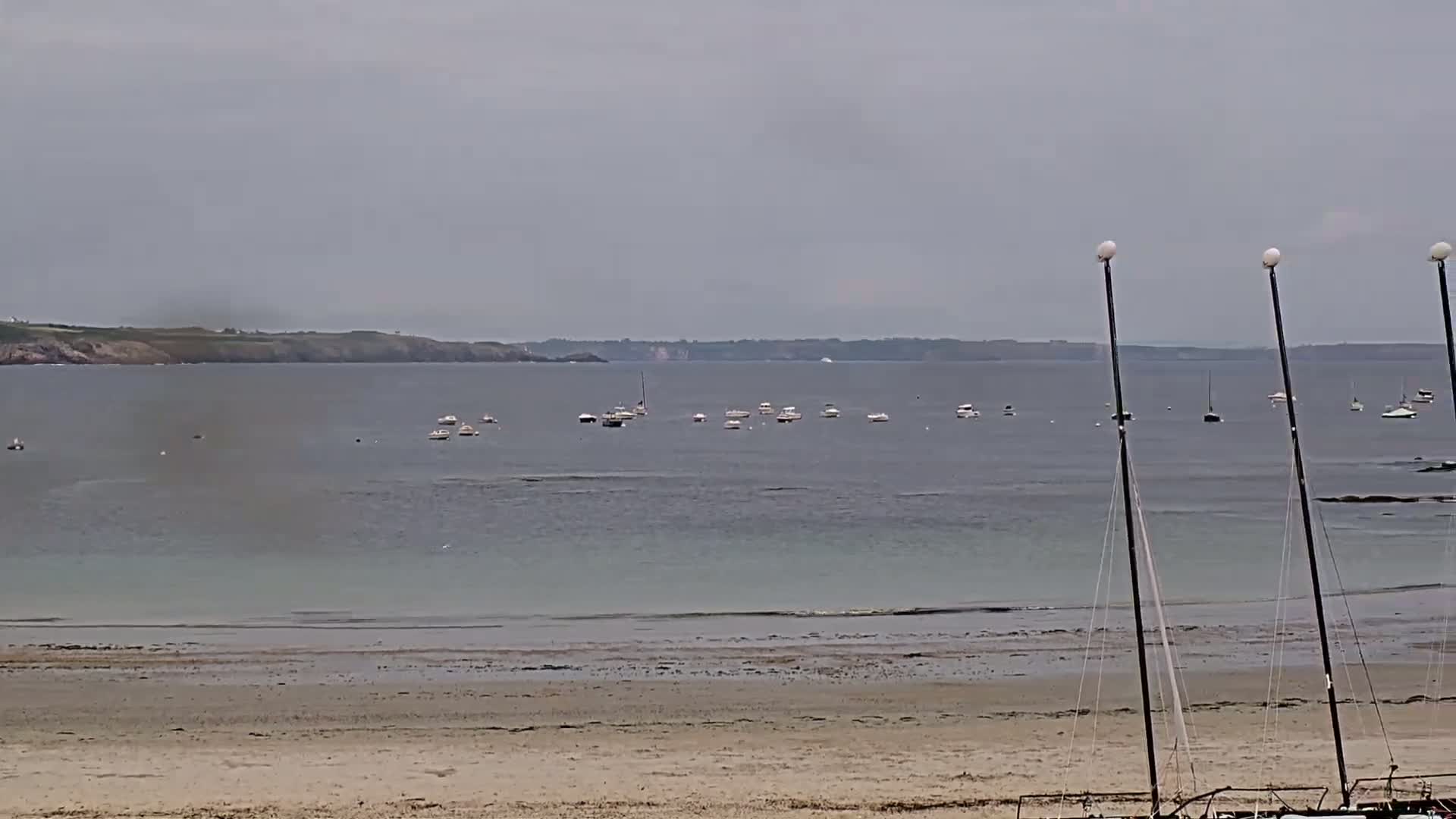 A sandy beach stretches to a calm, gray sea where several small boats are anchored; a distant shoreline is visible under an overcast sky.