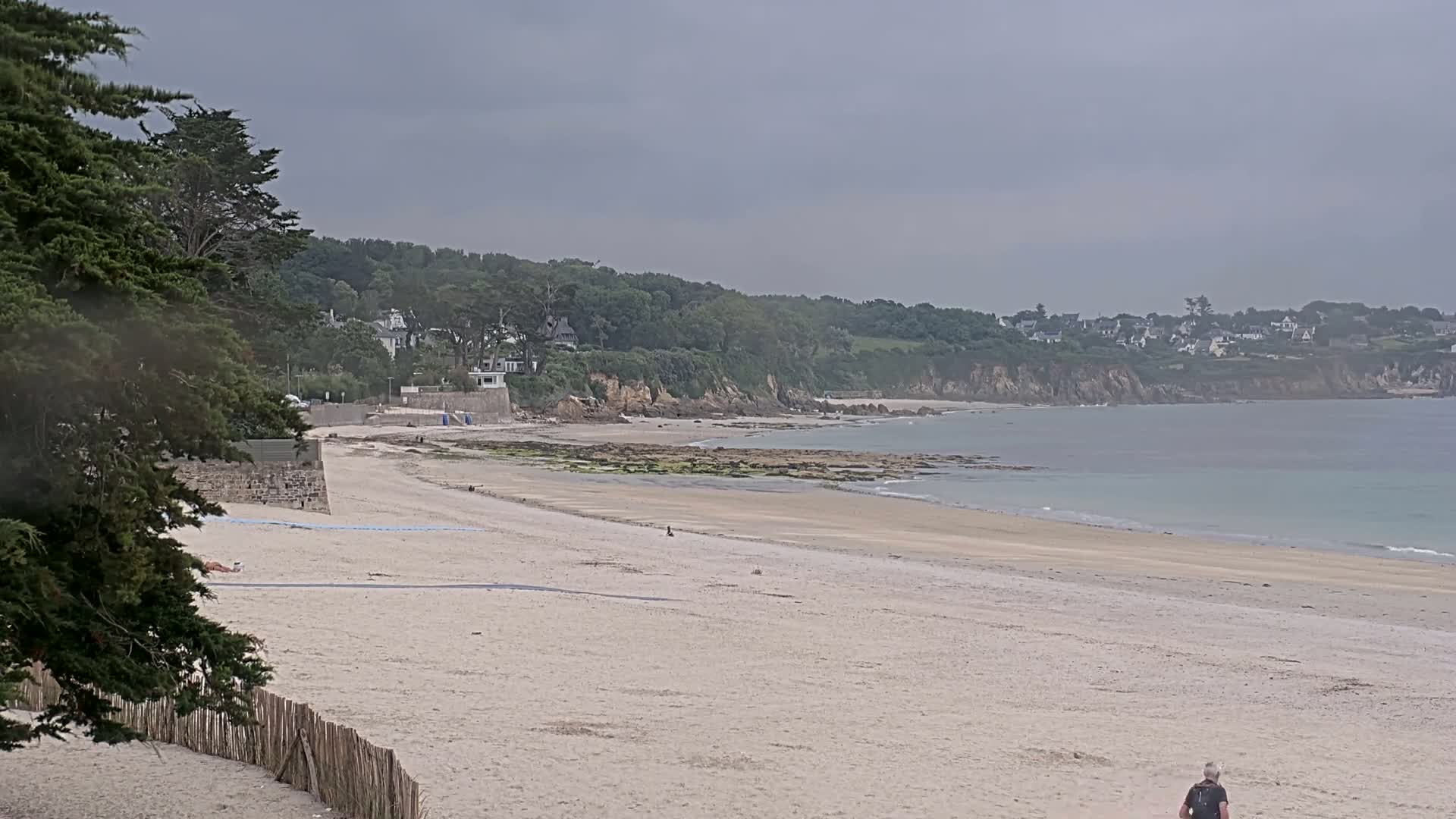 A sandy beach stretches out to a calm sea under an overcast sky, backed by a line of green cliffs and houses.