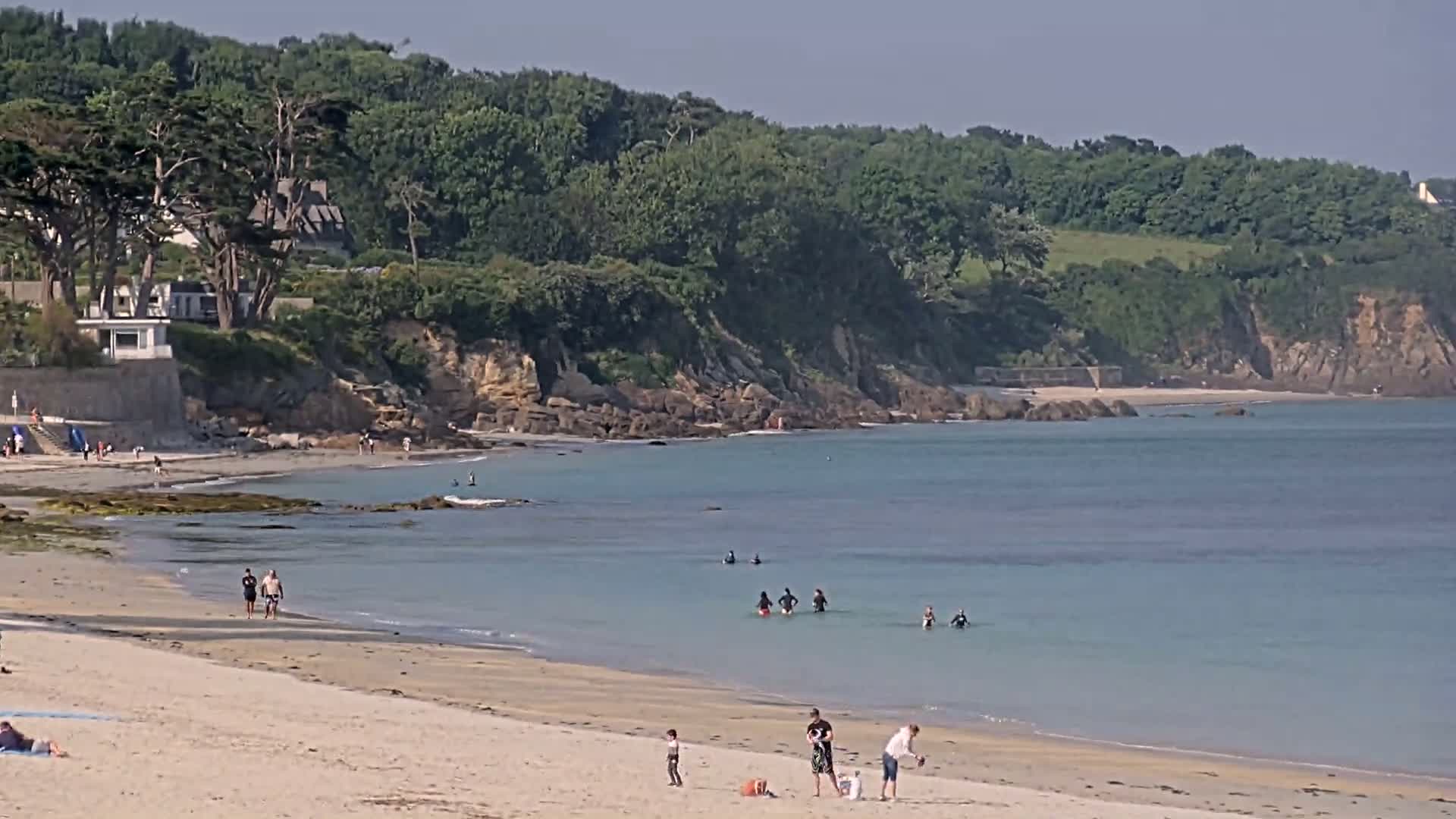 A sandy beach with calm, light-blue water, several people swimming and relaxing on the shore, backed by a green, tree-covered cliff on a sunny day.