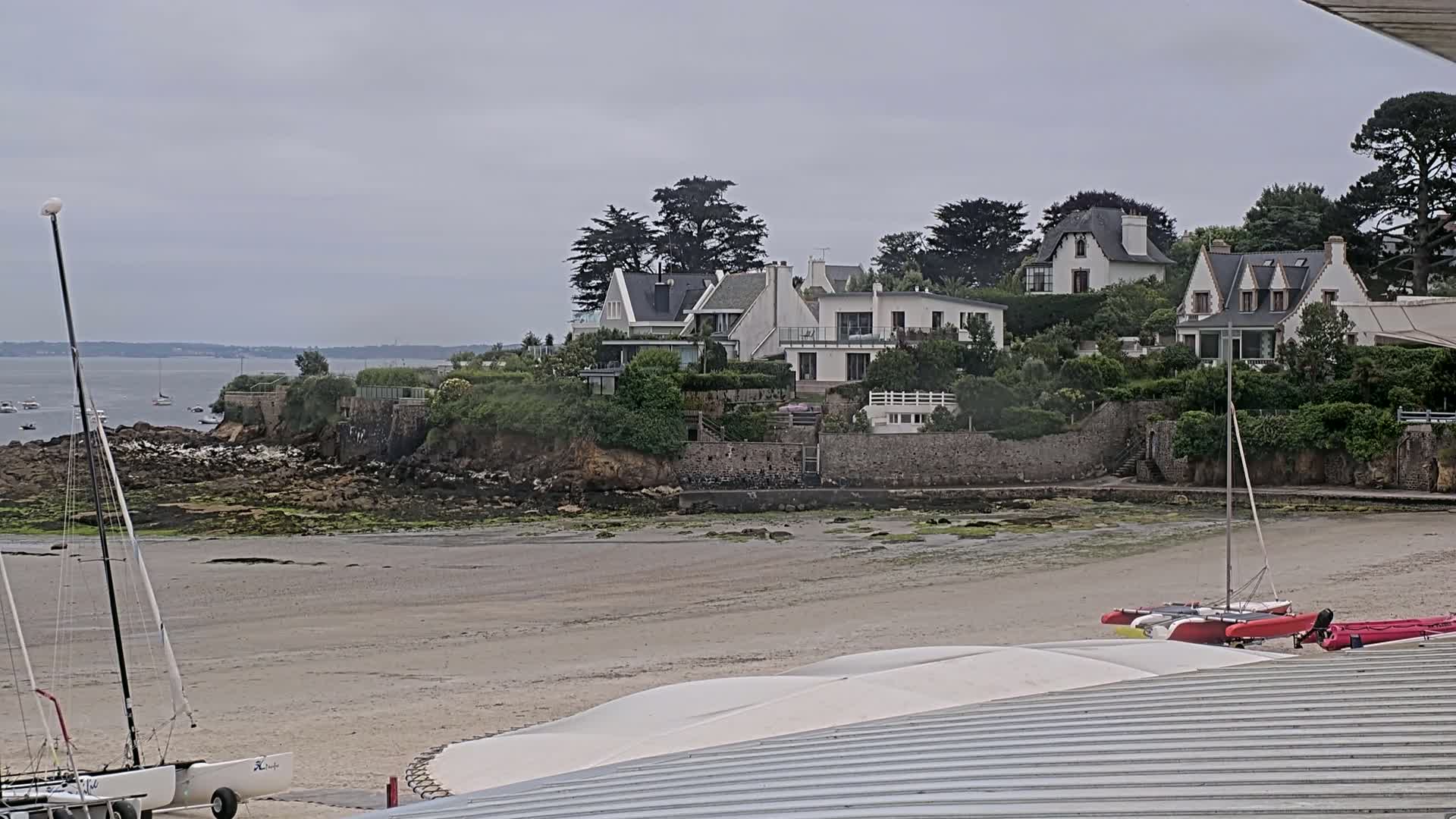 A sandy beach with several sailboats and a cluster of white houses on a green hillside overlooking calm water under an overcast sky.