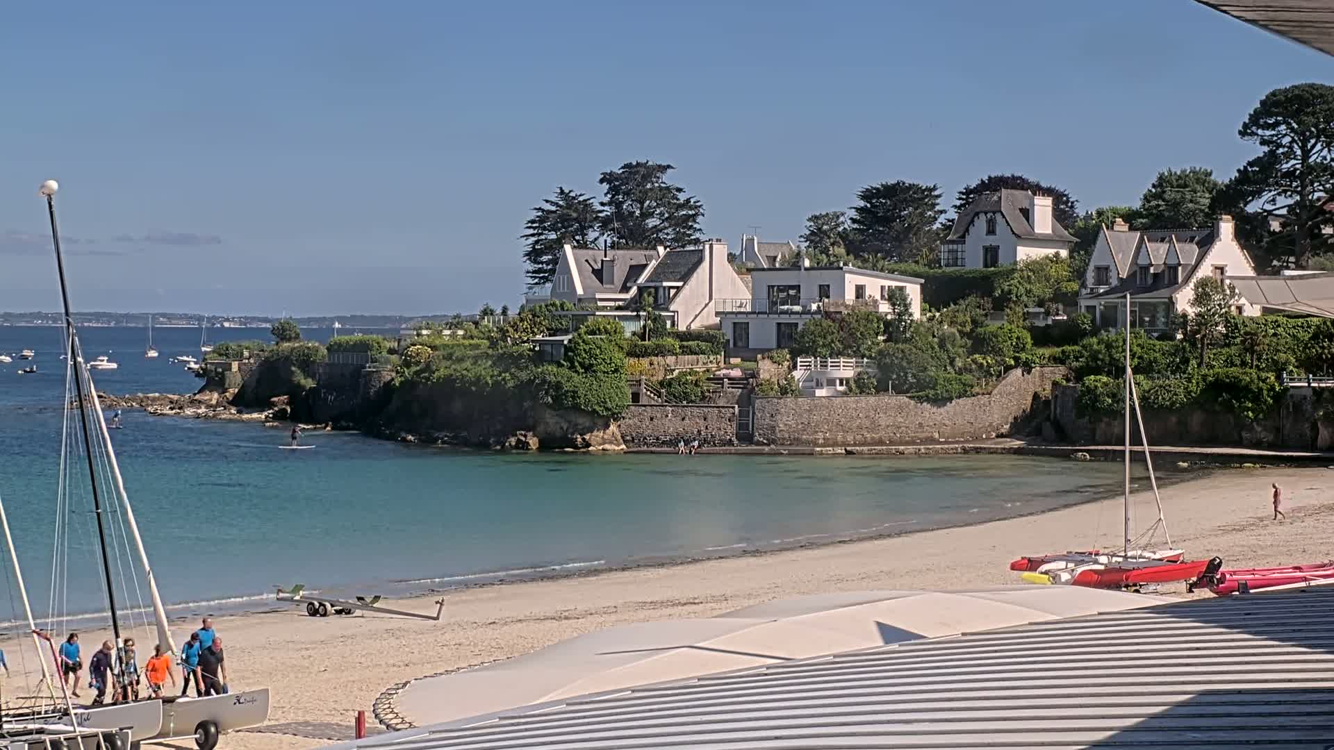 A sandy beach with several sailboats and people near the shore,  backed by a rocky outcrop and several white houses, under a clear blue sky.