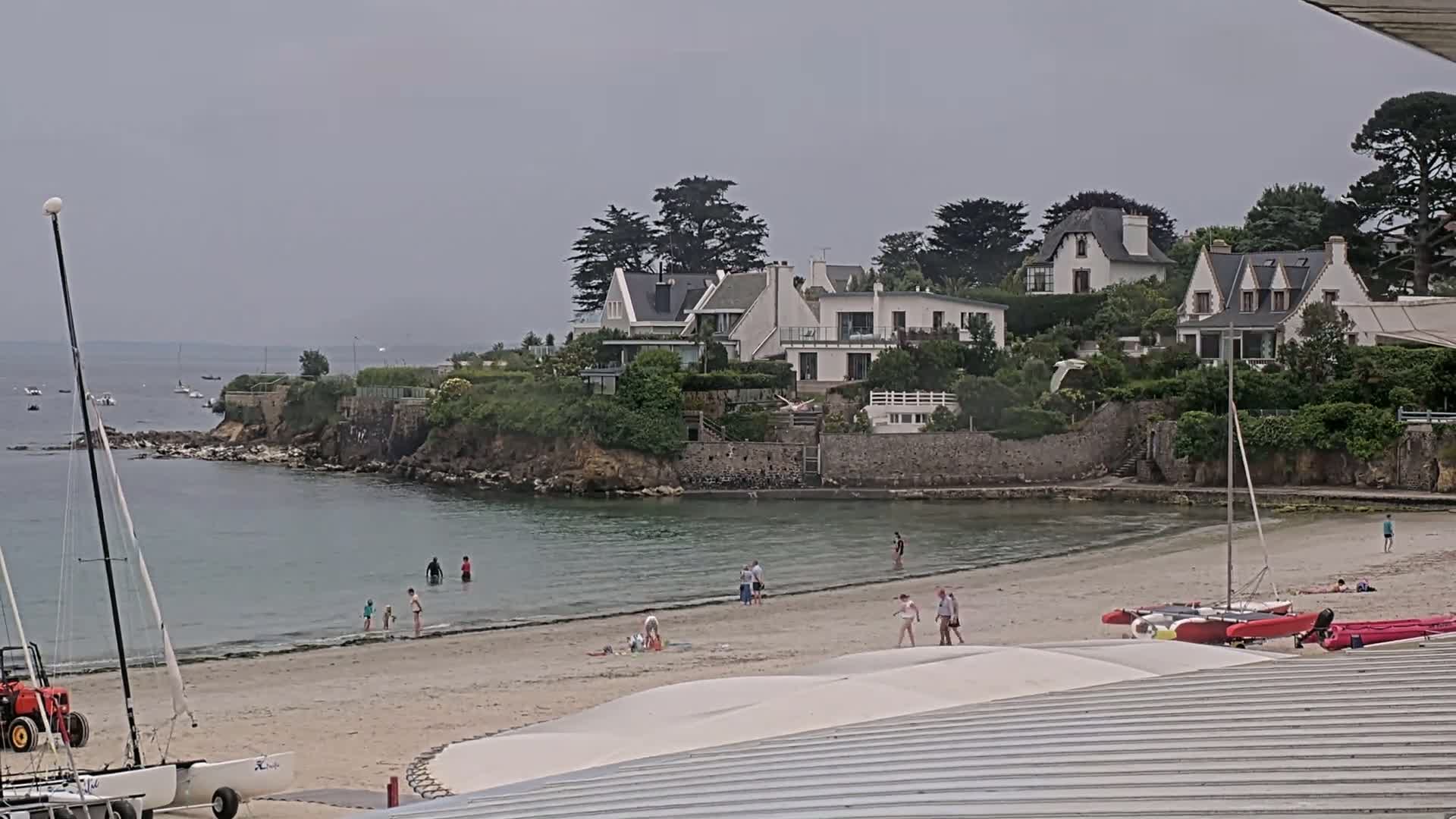 A sandy beach with several people swimming and relaxing, boats docked nearby, and white houses on a cliff overlooking a calm bay under an overcast sky.