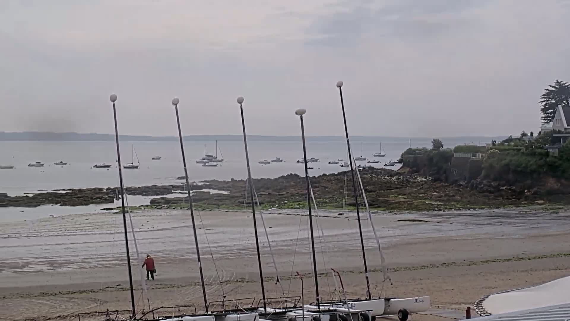 A sandy beach with several catamarans on the shore and numerous boats in the calm, overcast water beyond rocky intertidal zones.