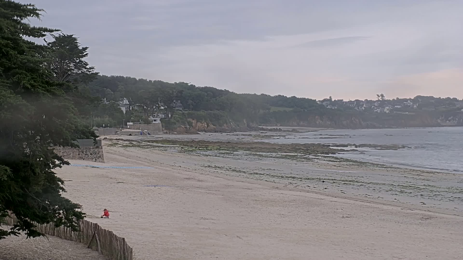 A sandy beach stretches out to a calm ocean under an overcast sky, with a few houses visible on a distant hillside.