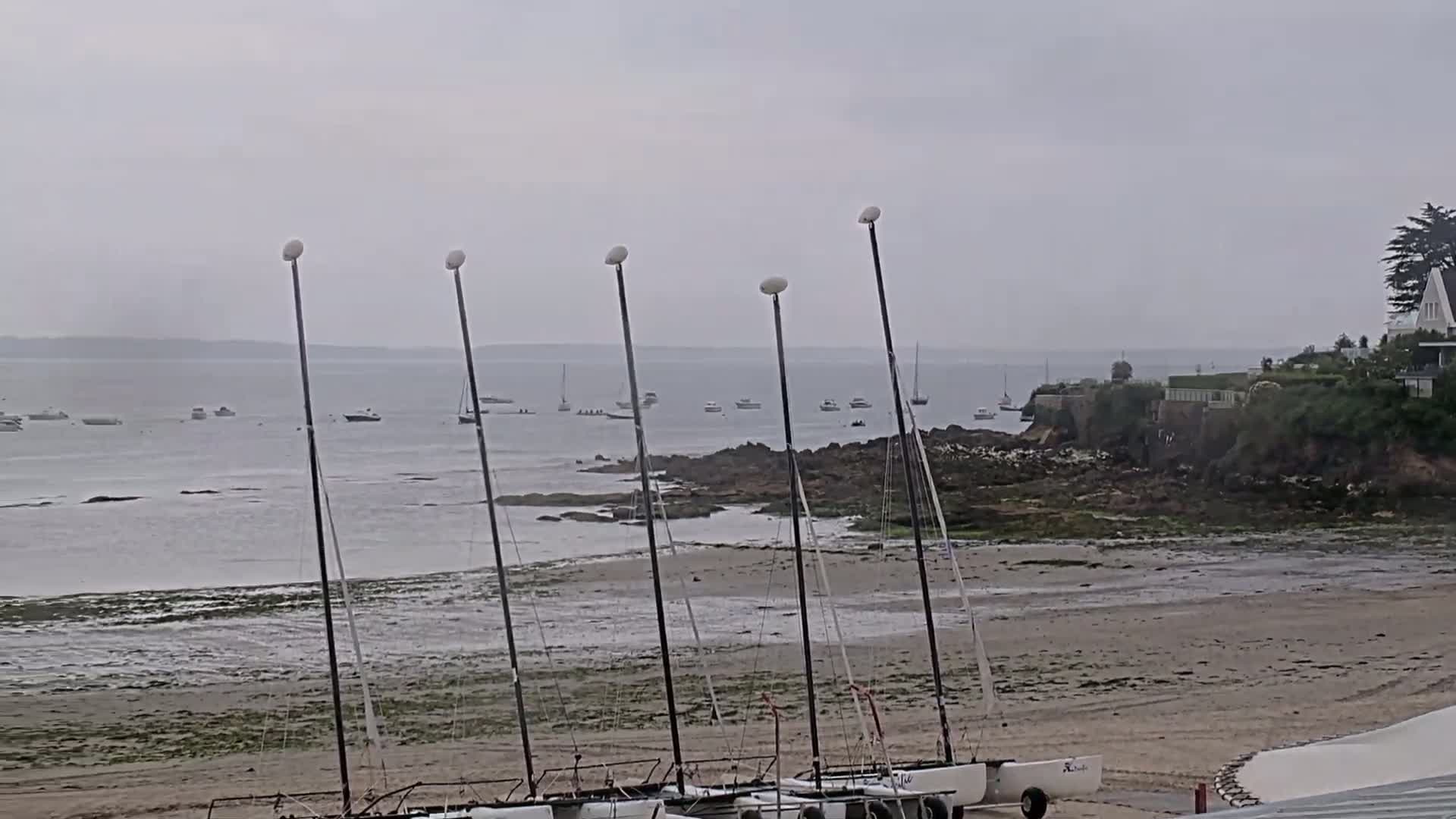 Several sailboats are beached on a sandy shore near rocky outcroppings, with a calm, overcast day visible in the background and several boats on the water.