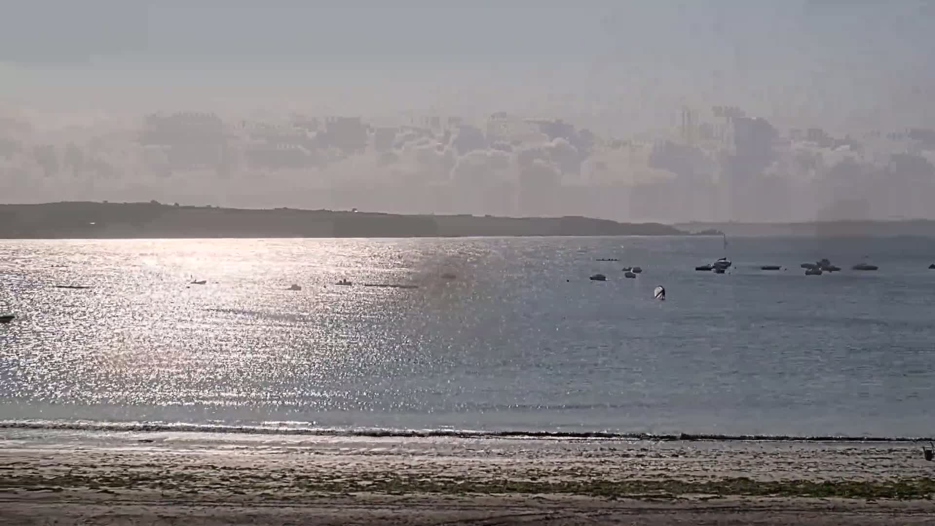 A sandy beach and calm, sunlit water with several small boats and a distant shoreline under a mostly cloudy sky.