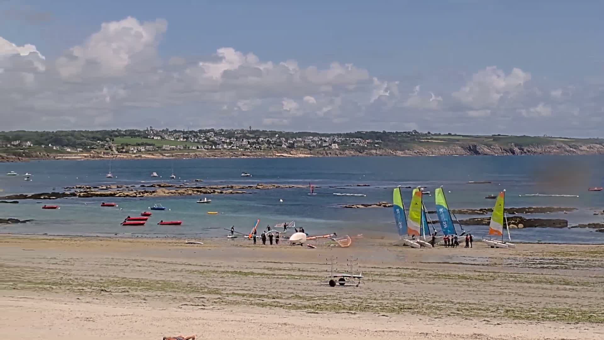 A sandy beach with several small sailboats beached near a group of people, under a partly cloudy sky, overlooking a calm bay with a town visible in the distance.