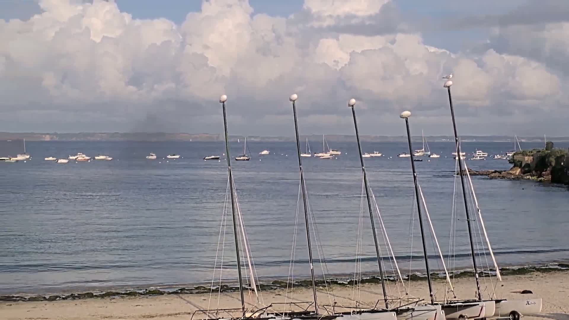 Several sailboats are beached on a sandy shore under a partly cloudy sky, with numerous other boats visible on the calm water in the distance.