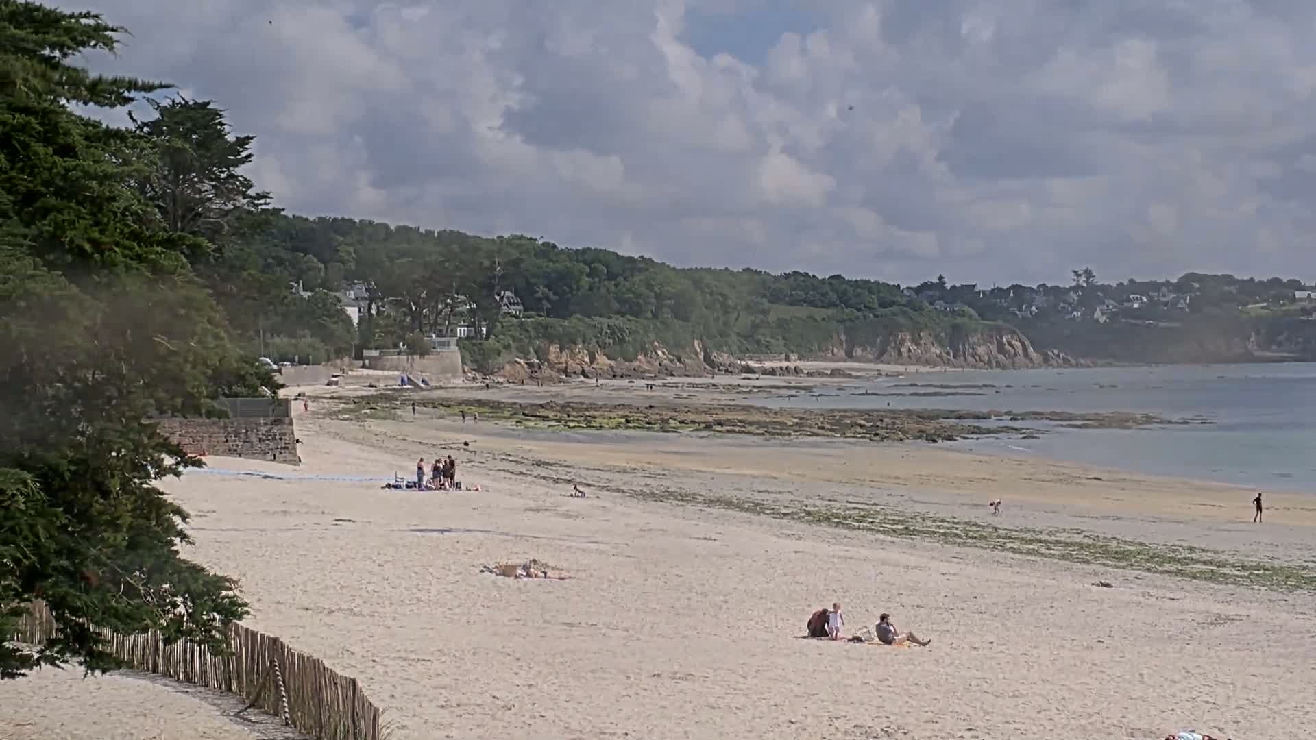 A sandy beach with a few people relaxing and some walking near the water's edge, under a partly cloudy sky, stretches out to a rocky shoreline and calm sea bordered by green hills and houses.