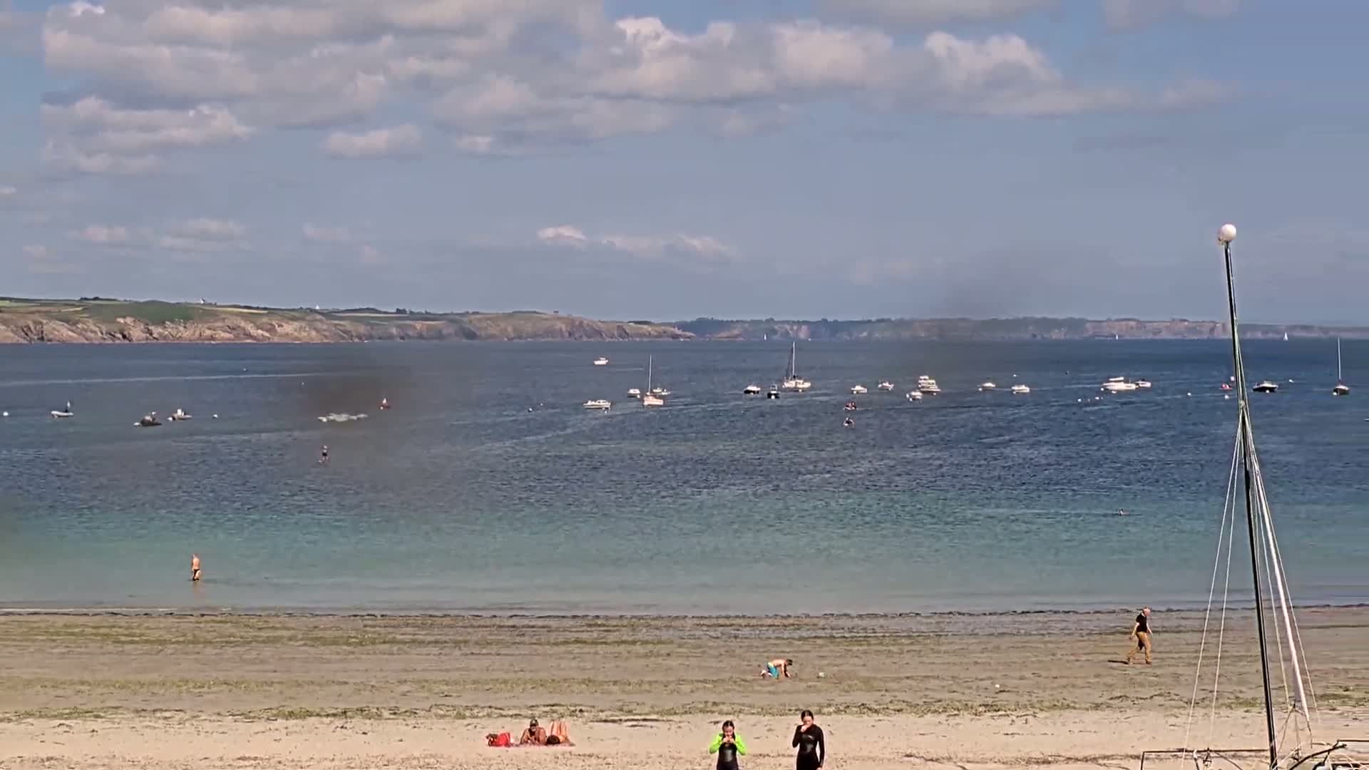 A sandy beach with several people on it and numerous boats in the calm, blue water under a partly cloudy sky.